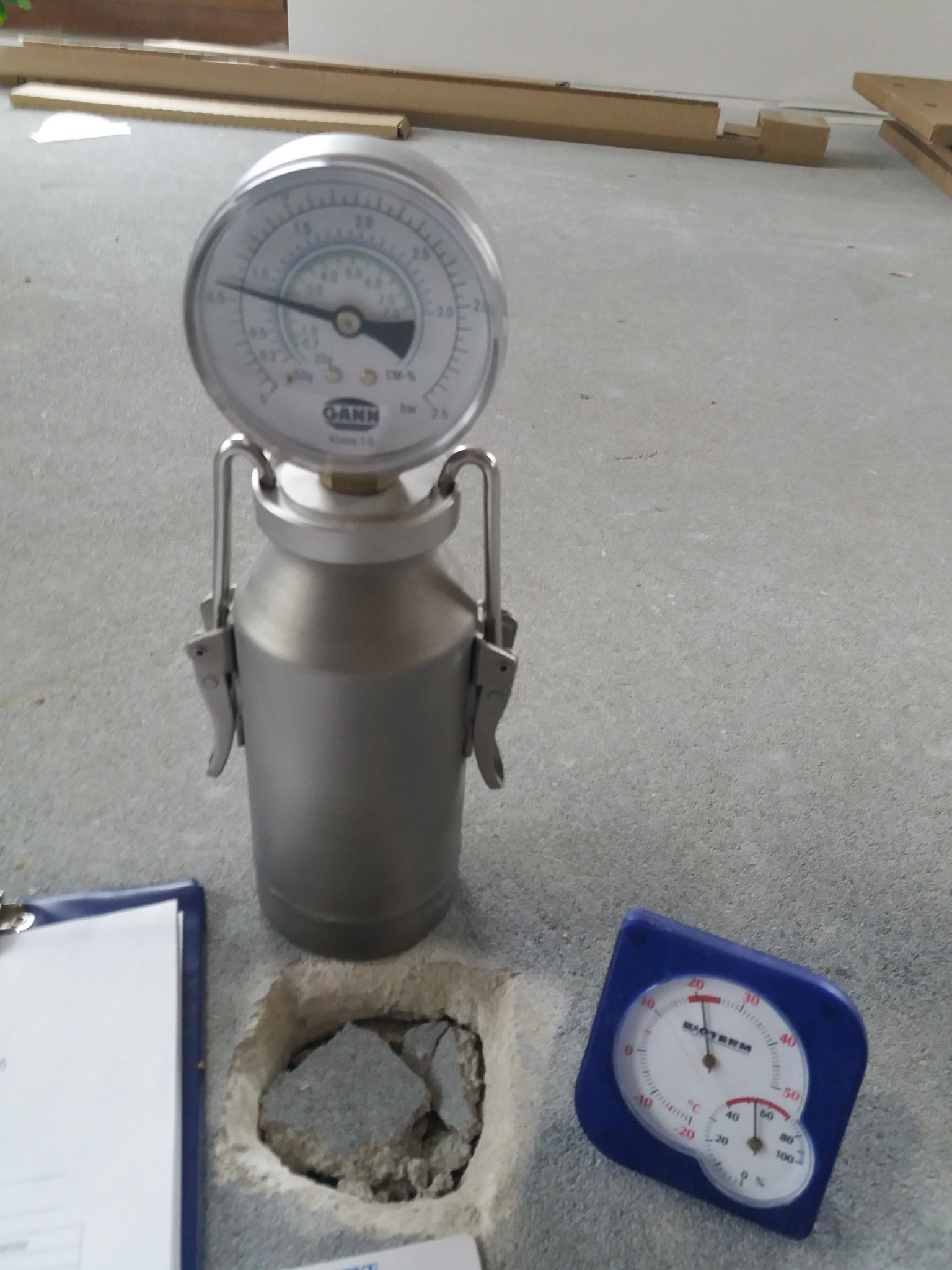 Pomiar wilgotności podłoża przed położeniem paneli: higrometr GANN na posadzce, obok termohigrometru Biorterm i notatnika. Widoczne kartony paneli w tle.