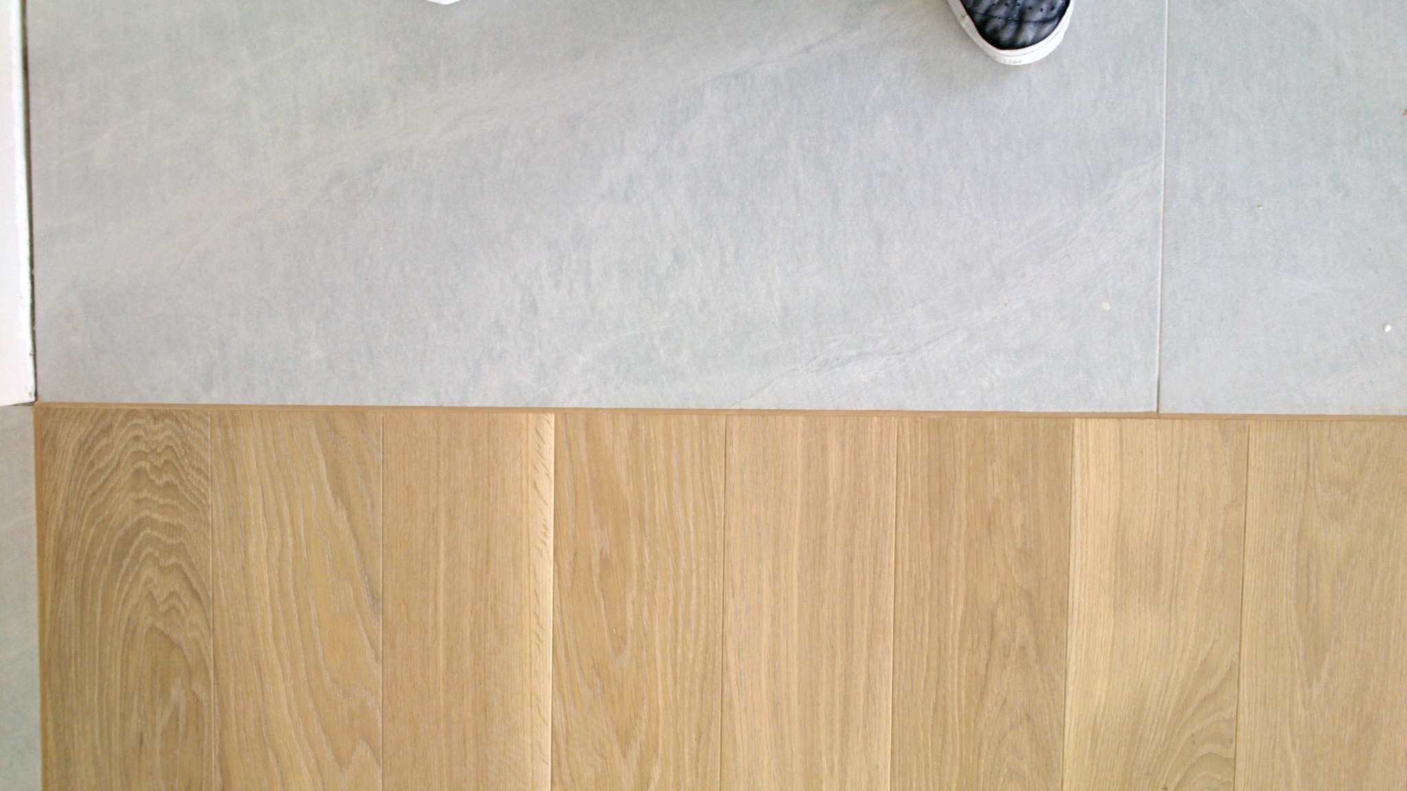 Połączenie jasnego parkietu z szarą kamienną posadzką, widok z góry. Fragment buta w rogu kadru. Naturalne światło.