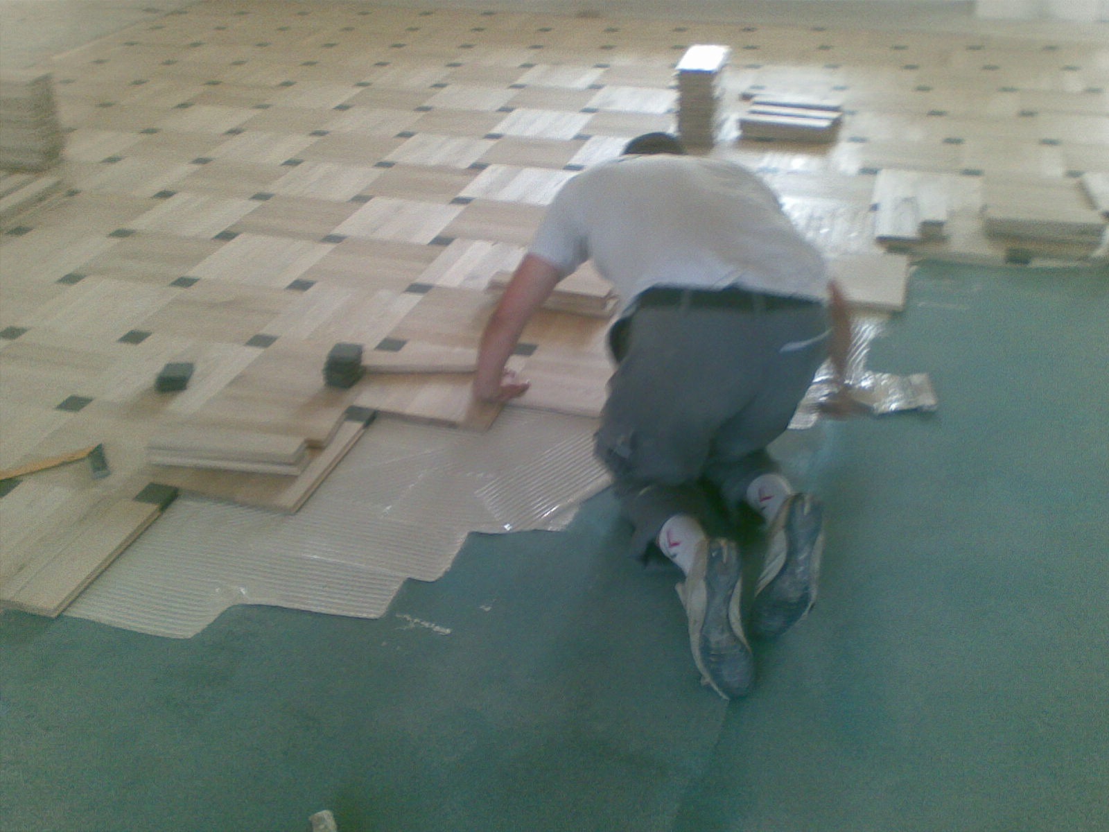 Pracownik na kolanach układa parkiet w geometryczny wzór, widoczne narzędzia i klej, fragment ukończonej podłogi. Charakterystyczne czarne akcenty we wzorze.