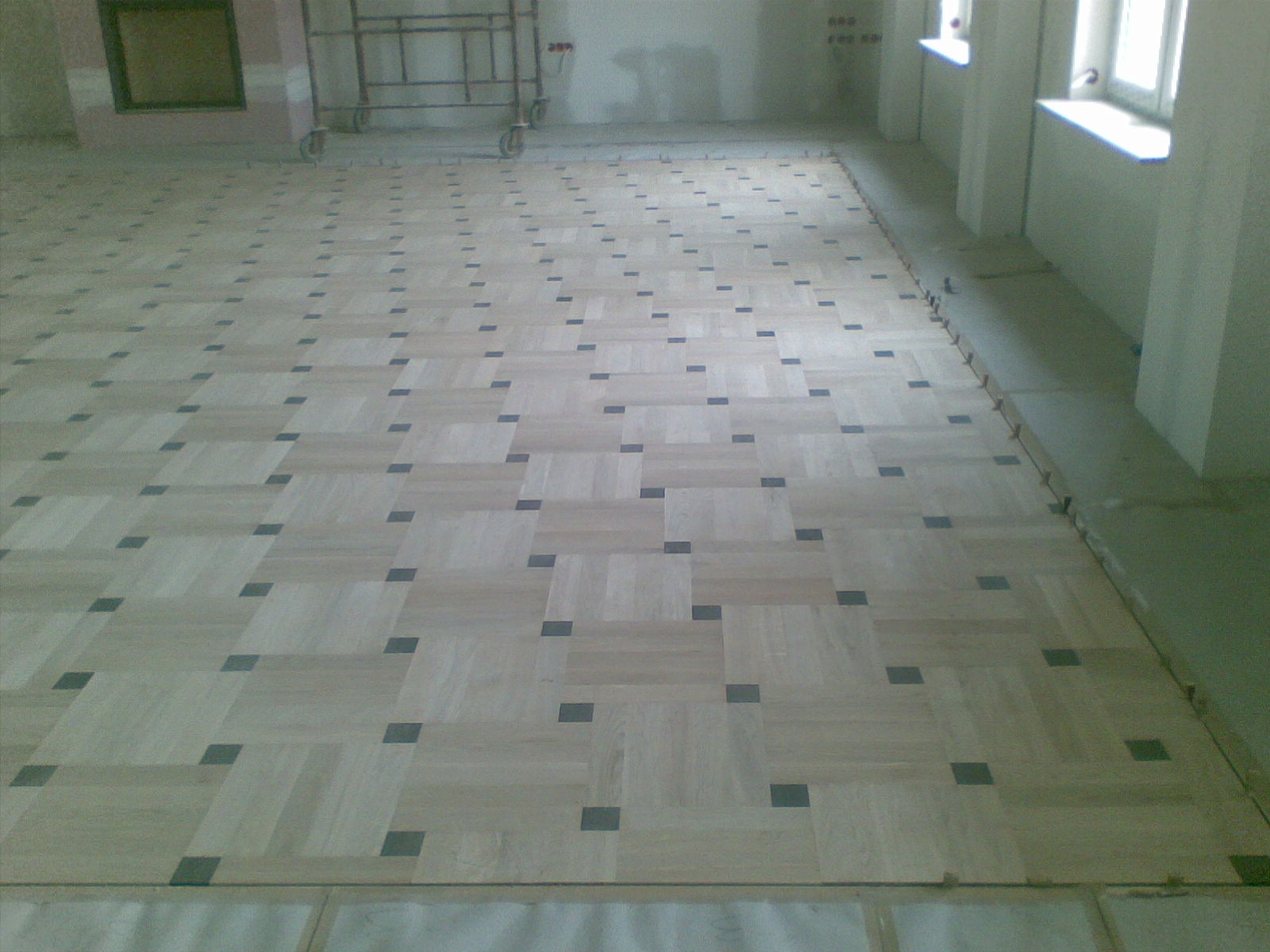 Jasny parkiet w kwadraty z ciemnymi akcentami, widok z góry na wykończoną podłogę w pustym pomieszczeniu z oknami i rusztowaniem w tle.