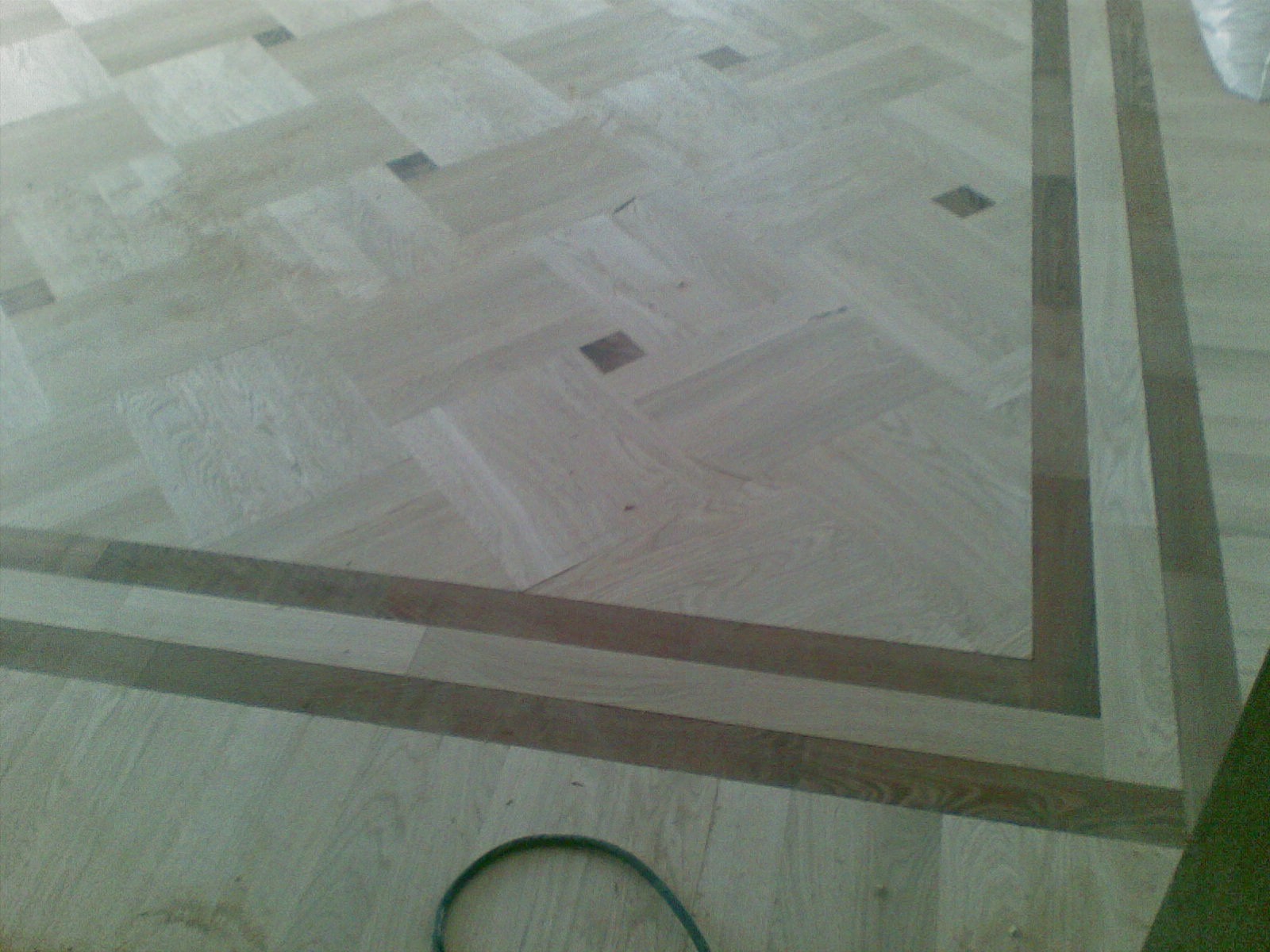 Jasny parkiet ułożony we wzór jodełki francuskiej z ciemnymi akcentami i bordiurą. Widoczny fragment kabla narzędzia w dolnej części kadru.