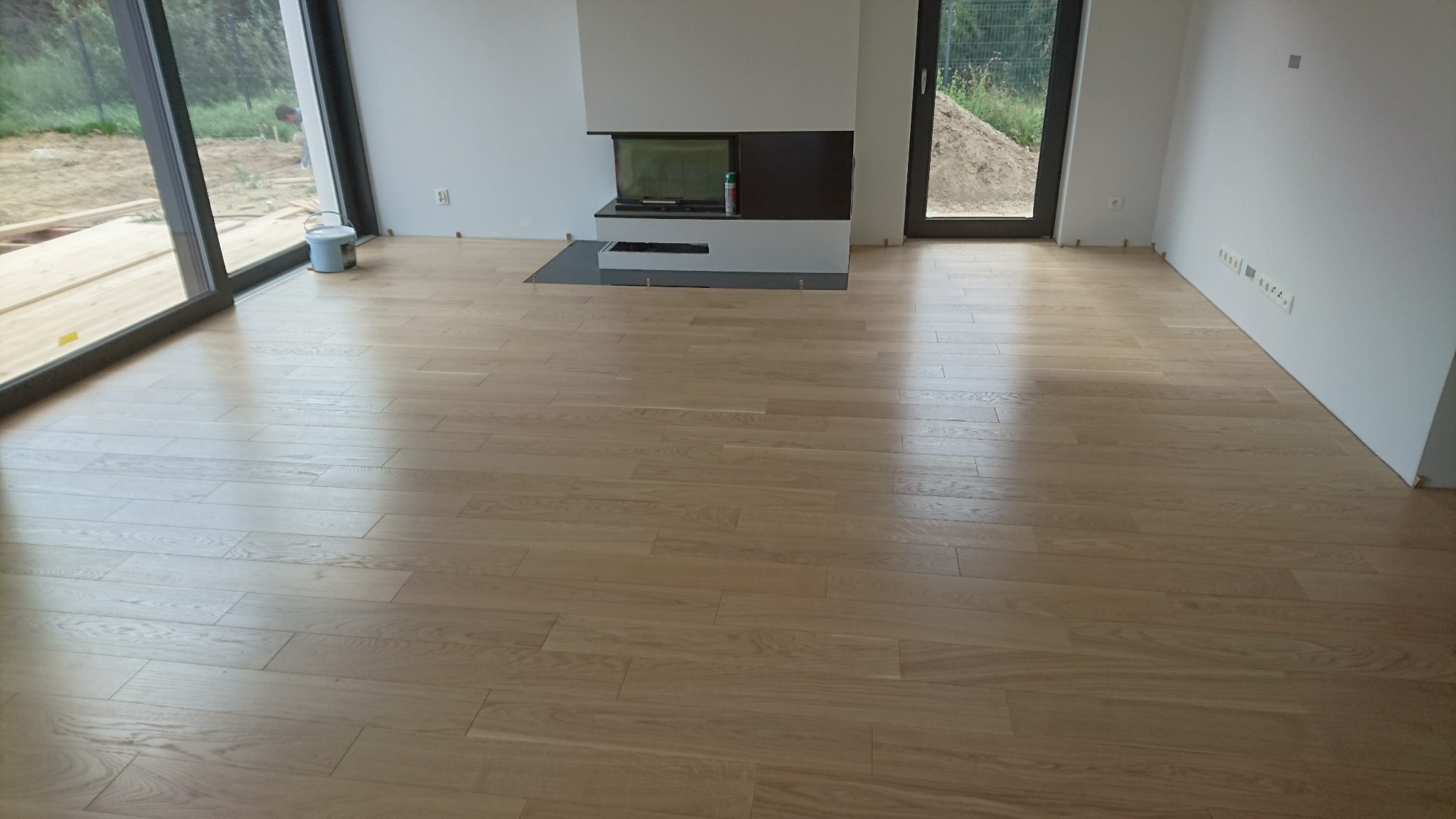 Jasny parkiet w nowoczesnym wnętrzu z kominkiem i dużymi oknami. Widoczny kubełek z farbą, świeżo położona podłoga, minimalistyczny design.