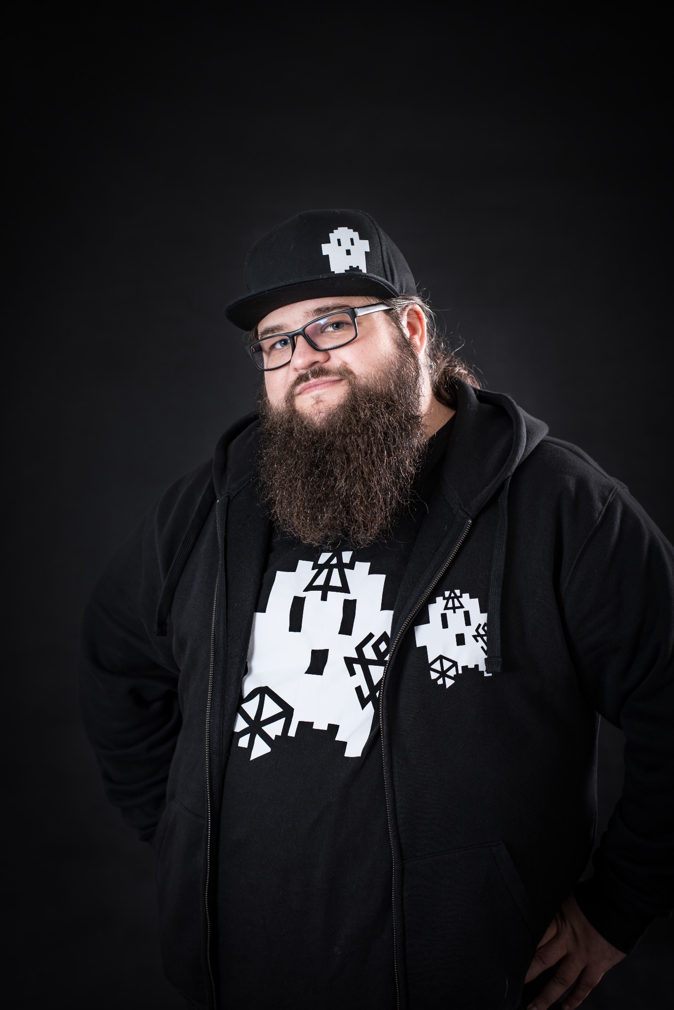 Mężczyzna z brodą w okularach, czarnej bluzie z kapturem i czapce z motywem pikselowego duszka na czarnym tle.