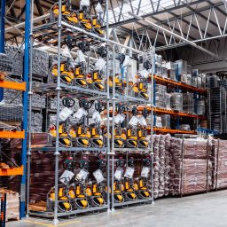 MegaMarket S. A. - Magazyn pełen nowych, żółto-czarnych wózków paletowych elektrycznych na metalowych regałach, widok z perspektywy podłogi, oświetlenie magazynowe.