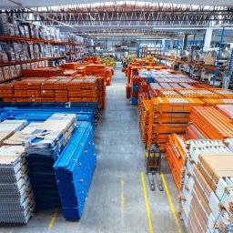 MegaMarket S. A. - Wysoki kąt widzenia na magazyn z wózkiem widłowym pomiędzy rzędami regałów wypełnionych pomarańczowymi, niebieskimi i srebrnymi elementami konstrukcyjnymi, widoczne żółte linie wyznaczające ścieżki.