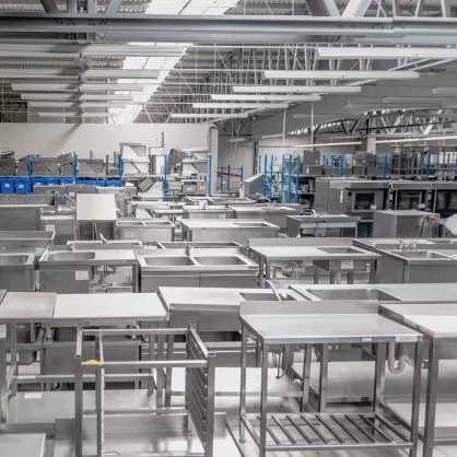 Magazyn pełen nowych, nierdzewnych stołów roboczych i zlewozmywaków gastronomicznych, widok z góry na halę produkcyjną.