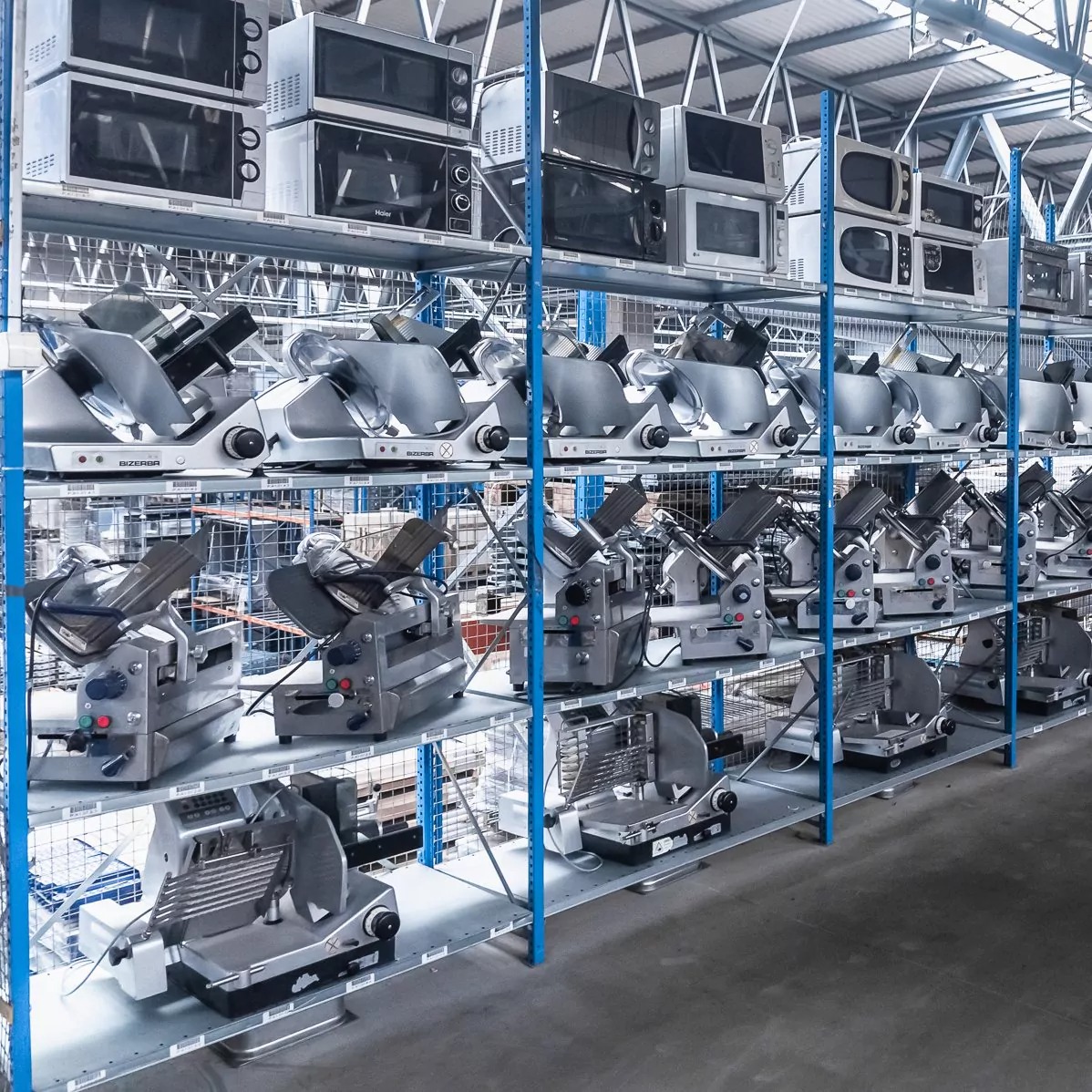 Magazyn z półkami, na których znajdują się krajalnice do wędlin Bizerba i kuchenki mikrofalowe, ustawione rzędami.