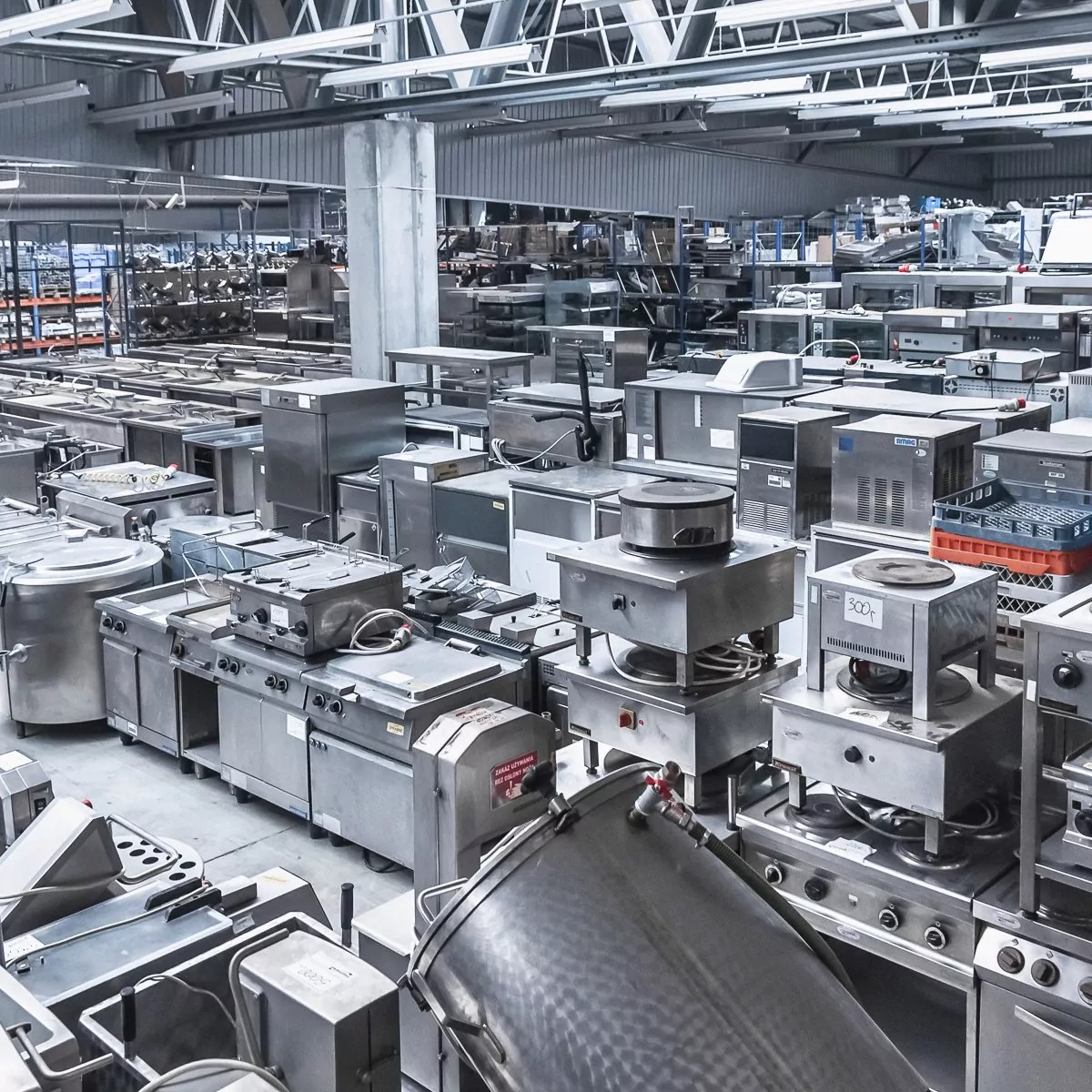 Magazyn pełen nowego, przemysłowego sprzętu kuchennego ze stali nierdzewnej, widok z góry, oświetlenie magazynowe, różnorodne urządzenia, w tym kuchenki, frytownice i pojemniki.
