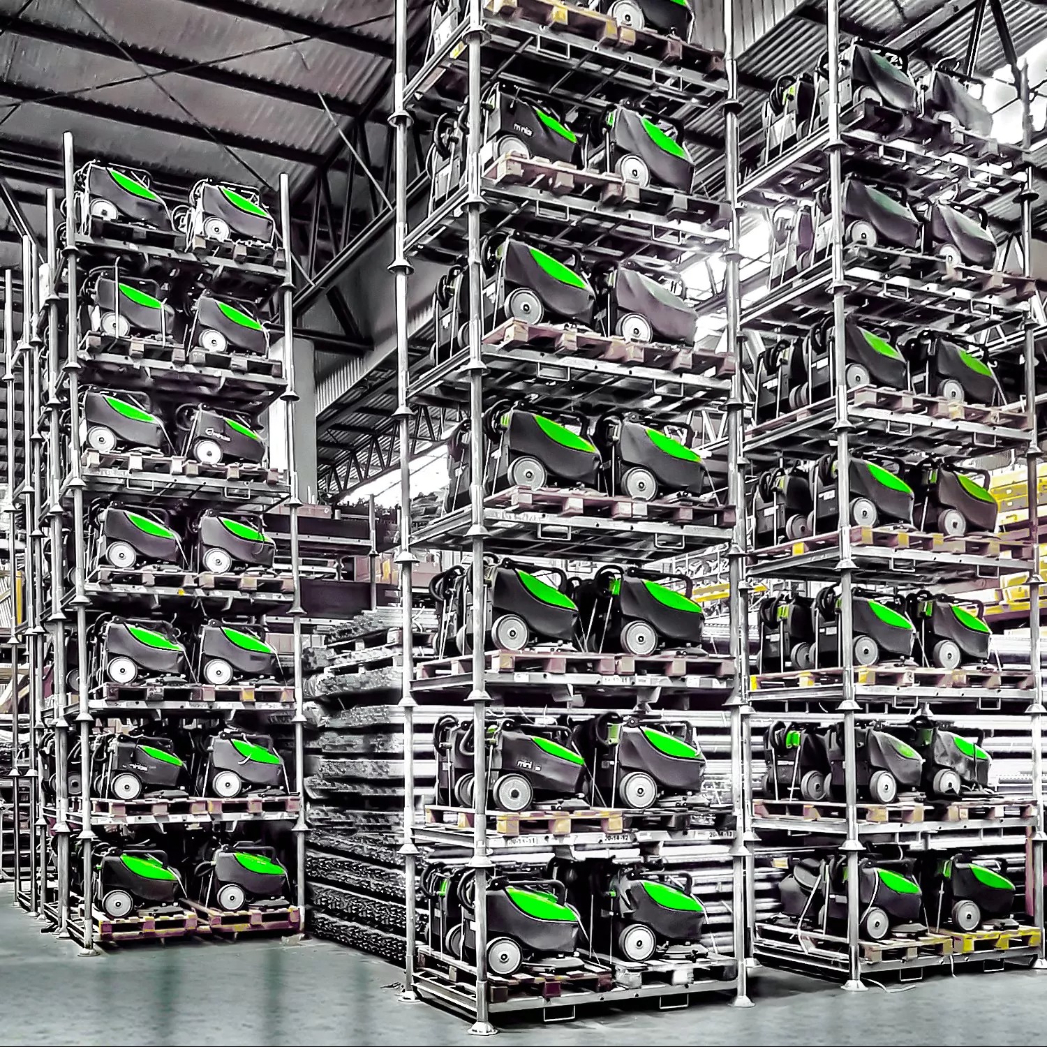 Magazyn pełen nowych, szarych wózków czyszczących z zielonymi akcentami, ustawionych rzędami na metalowych regałach.