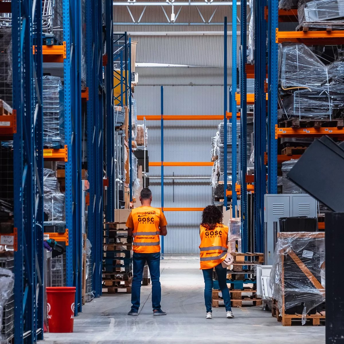Dwie osoby w pomarańczowych kamizelkach z napisem 'GOŚĆ VISITOR' stoją w magazynie między wysokimi regałami z niebieskimi słupkami i pomarańczowymi poprzeczkami, wypełnionymi towarami owiniętymi...