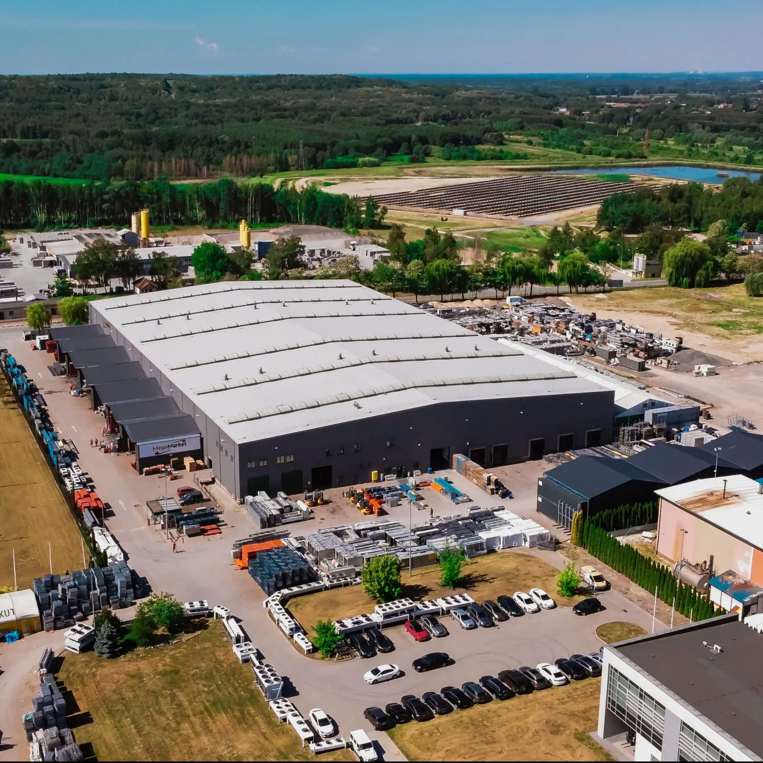 Szeroki plan magazynu MegaMarket z zaparkowanymi samochodami osobowymi, wózkami widłowymi i paletami na zewnątrz, otoczony zielenią i lasami w tle.