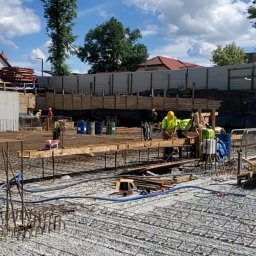 PERSHA VIZA SPÓŁKA Z OGRANICZONĄ ODPOWIEDZIALNOŚCIĄ - Prace zbrojarskie na budowie: widok na zbrojenie fundamentów z widocznymi pracownikami w żółtych kamizelkach, drewnianymi deskami i niebieskimi rurami na tle nieba z chmurami.