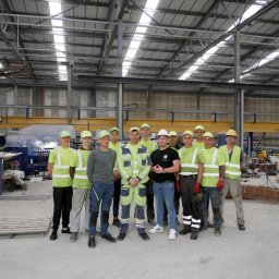 PERSHA VIZA SPÓŁKA Z OGRANICZONĄ ODPOWIEDZIALNOŚCIĄ - Grupa kilkunastu pracowników budowlanych w odblaskowych kamizelkach i czapkach, stojących w hali produkcyjnej na tle maszyn i elementów zbrojeniowych.