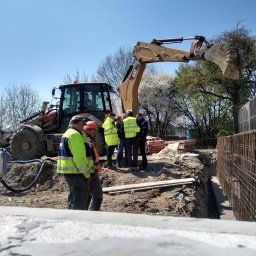 PERSHA VIZA SPÓŁKA Z OGRANICZONĄ ODPOWIEDZIALNOŚCIĄ - Prace ziemne w Oleśnicy: Grupa robotników w odblaskowych kamizelkach omawia projekt fundamentów przy wykopie i koparce CAT, widoczny szkielet zbrojenia.