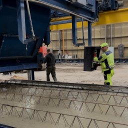 PERSHA VIZA SPÓŁKA Z OGRANICZONĄ ODPOWIEDZIALNOŚCIĄ - Pracownik w odblaskowym ubraniu obsługuje maszynę do wylewania betonu na konstrukcję zbrojeniową w hali produkcyjnej.