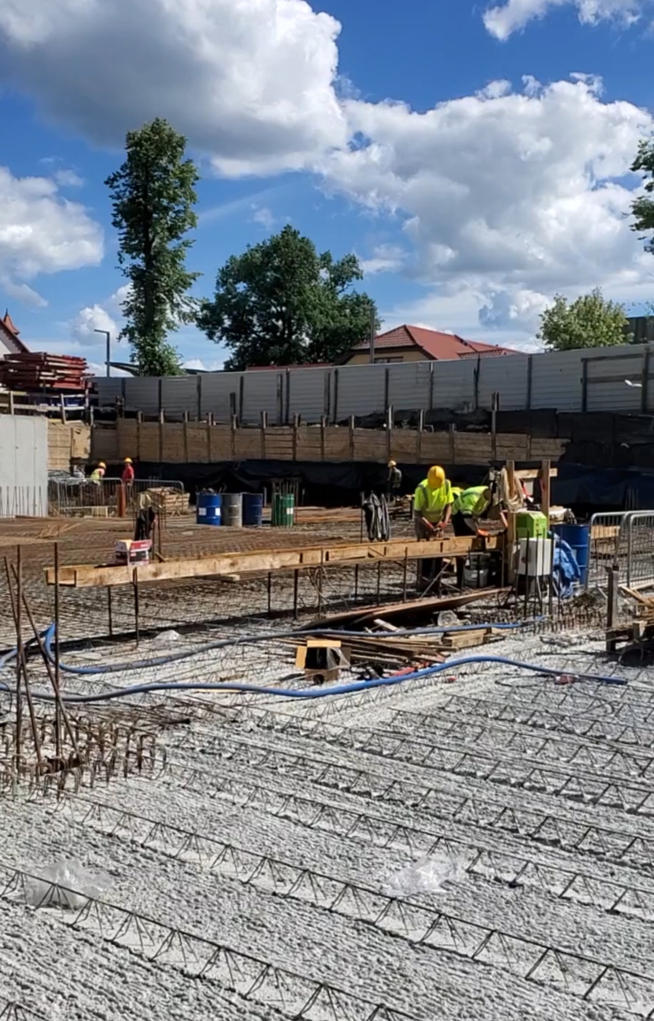 Prace zbrojarskie na budowie: widok na zbrojenie fundamentów z widocznymi pracownikami w żółtych kamizelkach, drewnianymi deskami i niebieskimi rurami na tle nieba z chmurami.