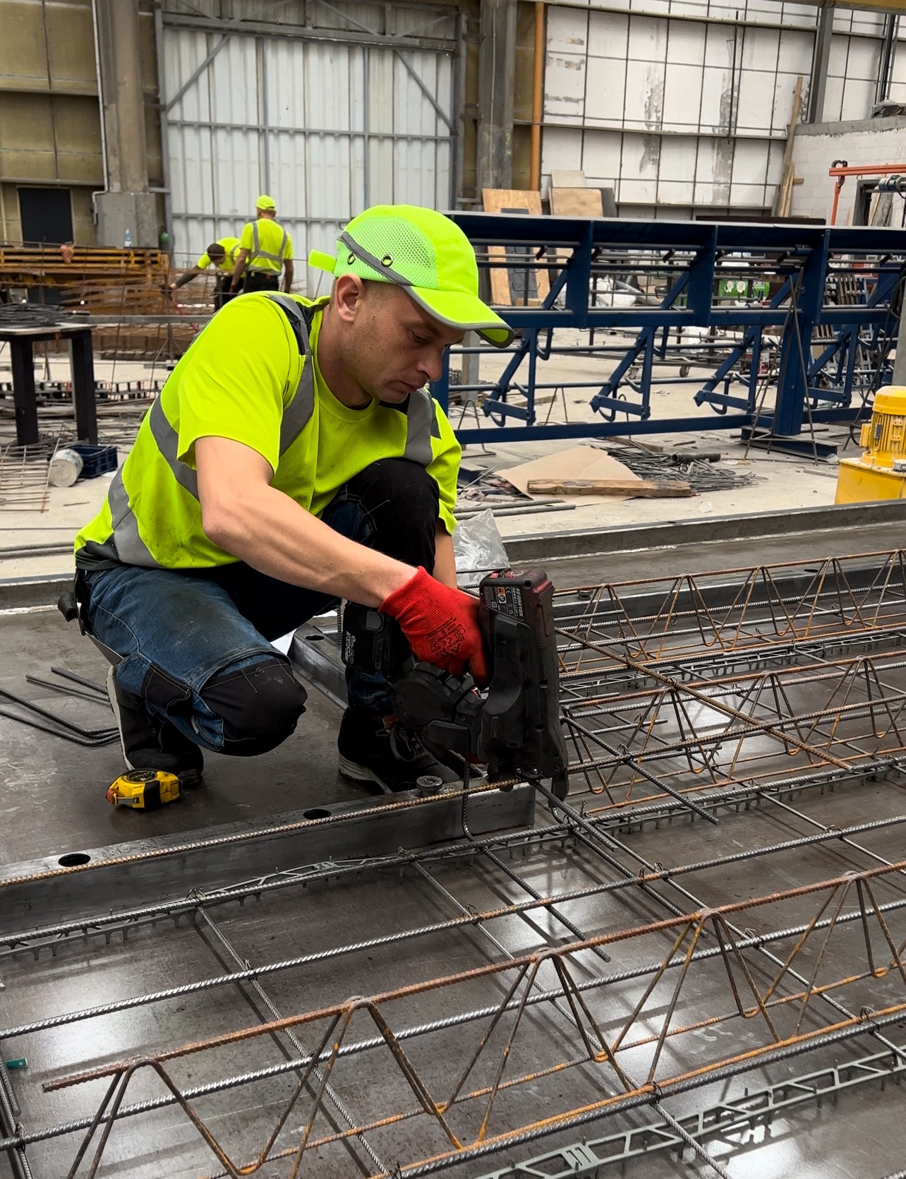 Pracownik w odblaskowej odzieży i czerwonych rękawicach łączy zbrojenie za pomocą specjalistycznego narzędzia na metalowej płycie; w tle hala produkcyjna z innymi pracownikami i konstrukcjami.
