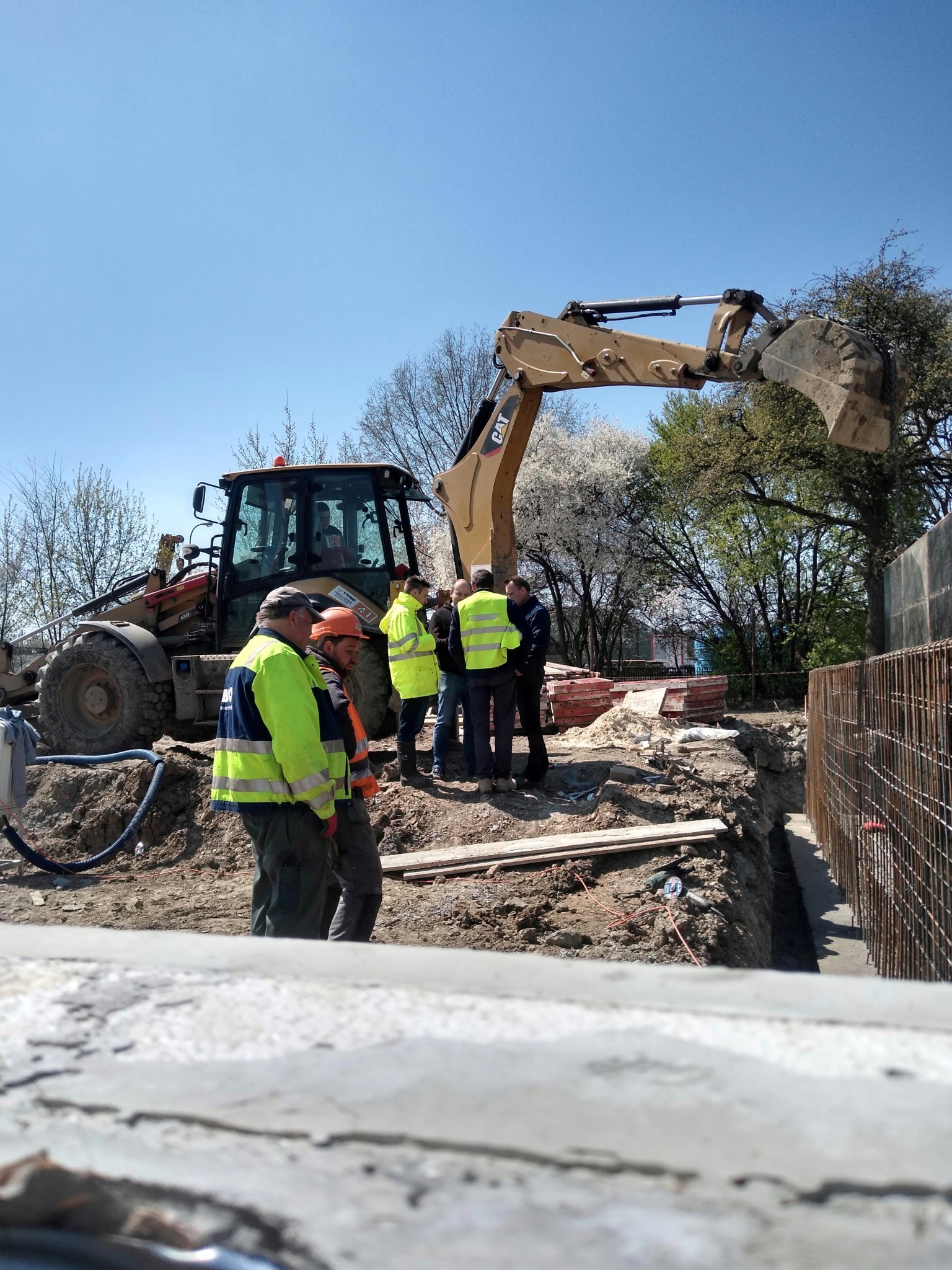 Prace ziemne w Oleśnicy: Grupa robotników w odblaskowych kamizelkach omawia projekt fundamentów przy wykopie i koparce CAT, widoczny szkielet zbrojenia.