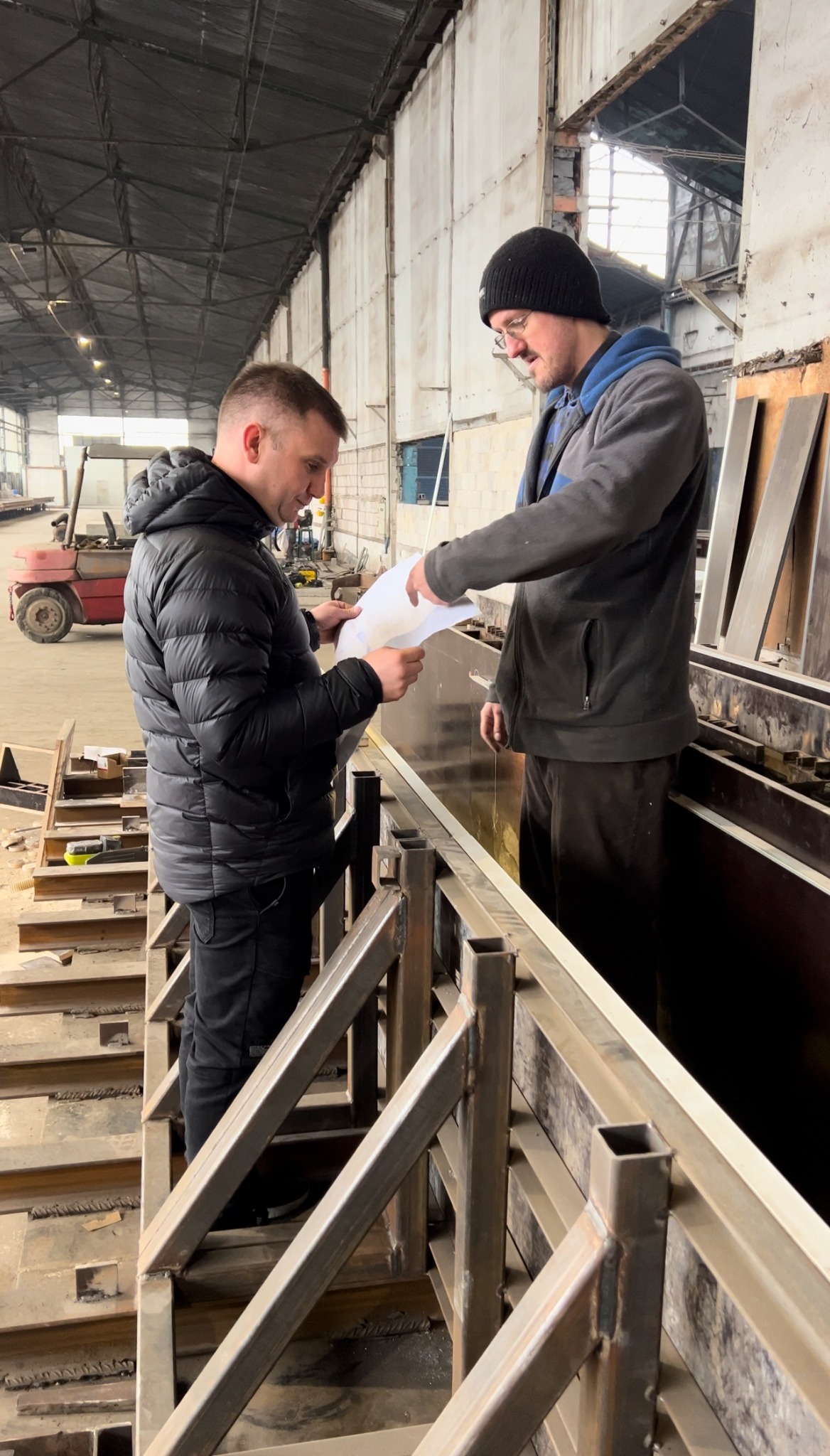 Dwóch mężczyzn w hali produkcyjnej omawia projekt na papierze, stojąc obok metalowej konstrukcji przypominającej szalunek, z widocznym w tle wózkiem widłowym.