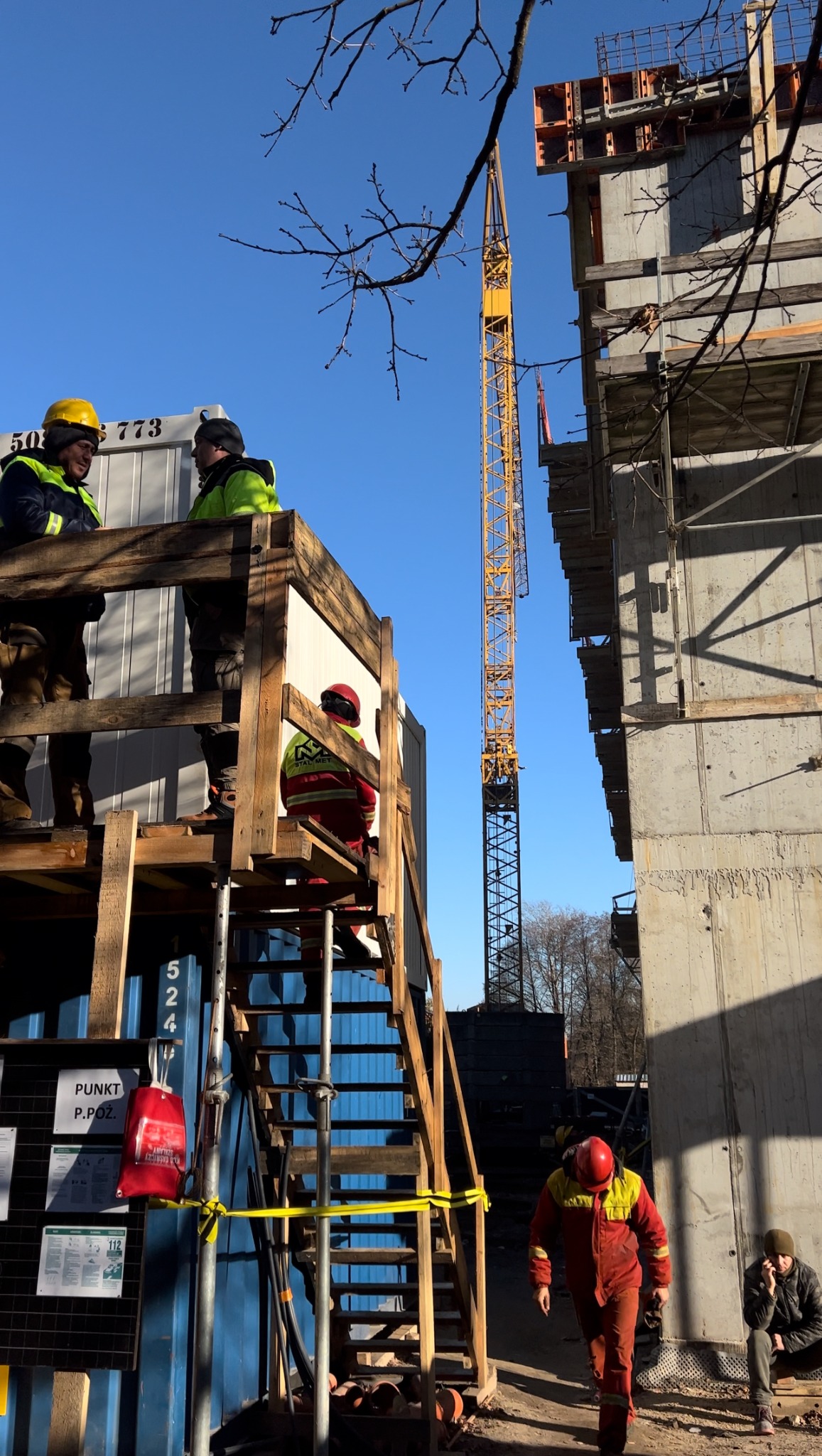 Żuraw budowlany góruje nad placem budowy, gdzie pracownicy w kaskach i kombinezonach poruszają się po drewnianych rusztowaniach i schodach, a betonowa konstrukcja budynku z widocznym szalunkiem...
