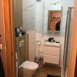 TOMDOM - Ukończona łazienka z białymi marmurkowymi płytkami, drewnianymi akcentami, wiszącą toaletą, białą szafką z umywalką i kabiną prysznicową. Widoczne odbicie osoby w lustrze.