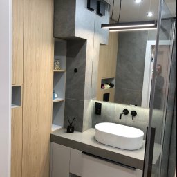 TOMDOM - Nowoczesna łazienka z drewnianą szafką, szarą umywalką nablatową i czarną armaturą, oświetlona podłużną lampą LED, z widocznym fragmentem kabiny prysznicowej.