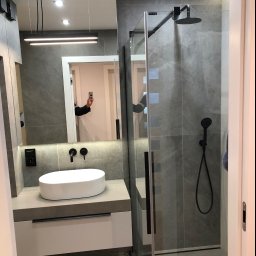 TOMDOM - Nowoczesna łazienka z szarymi płytkami, kabiną prysznicową typu walk-in z czarną armaturą, owalną białą umywalką nablatową i podświetlanym lustrem, w którym odbija się osoba robiąca zdjęcie telefonem.