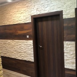 TOMDOM - Wnętrze z dekoracyjną ścianą z imitacji kamienia w odcieniach beżu, przedzieloną poziomym pasem z paneli drewnopodobnych w kolorze ciemnego brązu, oraz z ciemnymi drzwiami z metalową klamką.