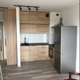 TOMDOM - Nowoczesna kuchnia z drewnianymi szafkami, czarnym blatem i marmurowym backsplashiem, oświetlona reflektorami sufitowymi, z widoczną lodówką i fragmentem podłogi z paneli imitujących drewno.