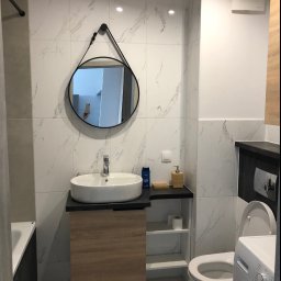 TOMDOM - Nowoczesna łazienka z marmurowymi płytkami, owalną umywalką na szafce z drewnopodobnym frontem, okrągłym lustrem na pasku i pralką obok toalety.