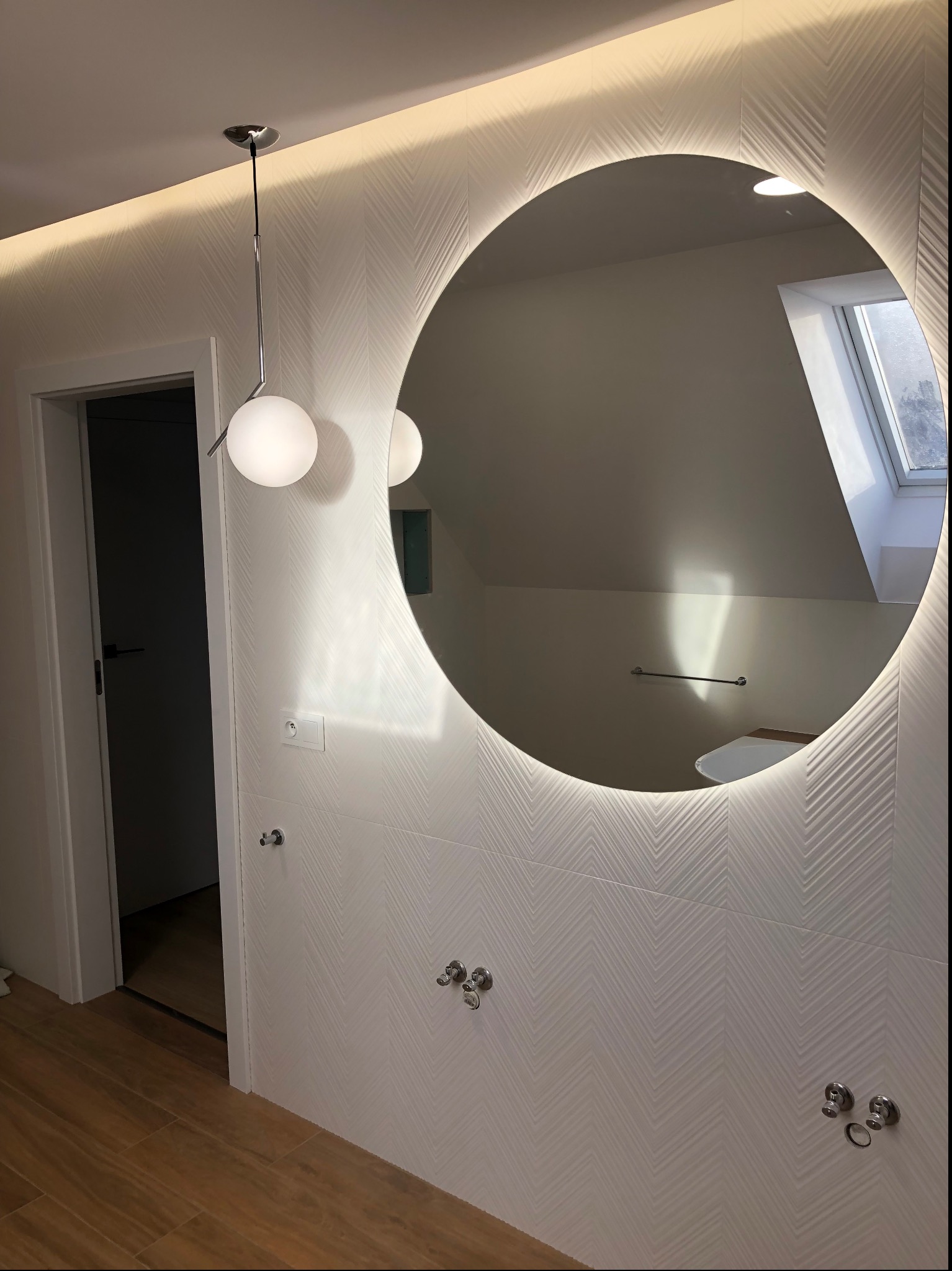 Nowoczesna łazienka z okrągłym lustrem podświetlanym LED, białe płytki z subtelnym wzorem, drewnopodobna podłoga, wisząca lampa z kulistym kloszem i widoczne elementy armatury