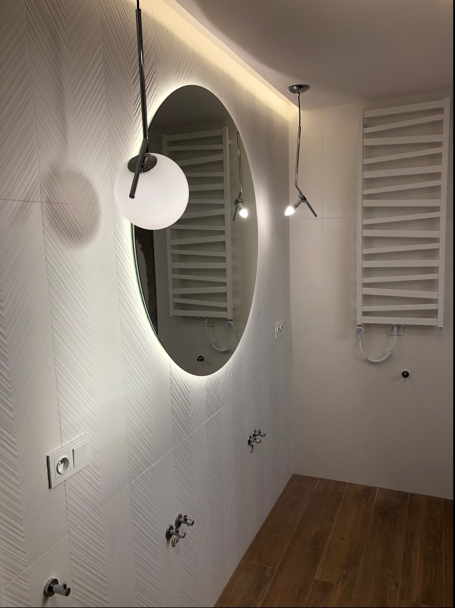 Nowoczesna łazienka z owalnym lustrem podświetlonym LED, designerskim oświetleniem wiszącym i grzejnikiem drabinkowym na ścianie z wzorzystych, białych płytek, uzupełnona drewnopodobną podłogą
