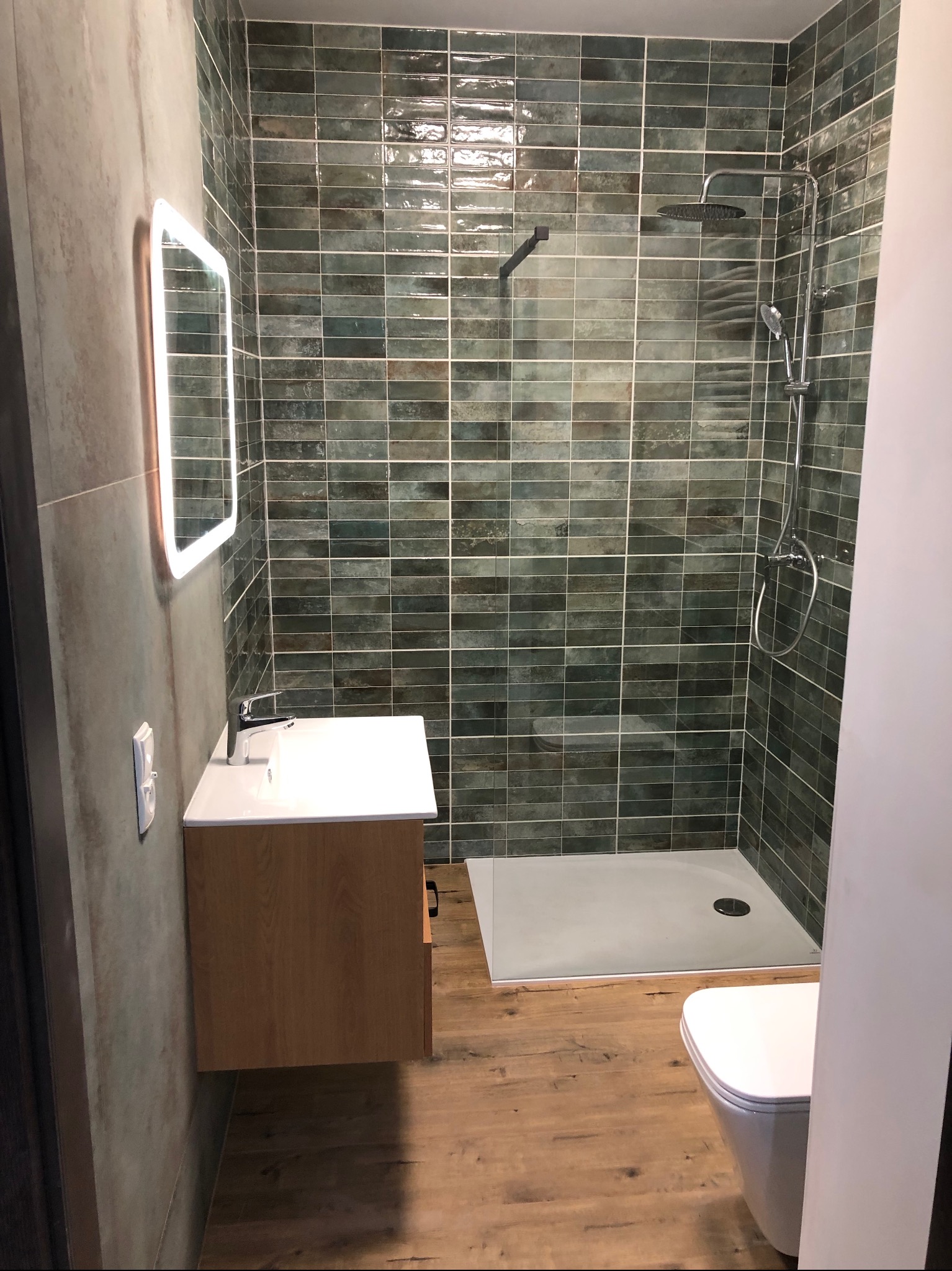 Nowoczesna łazienka z kabiną prysznicową typu walk-in, umywalką na szafce z drewnianym fornirem i podświetlanym lustrem LED, wykończona płytkami w odcieniach zieleni i brązu, z drewnopodobną podłogą.