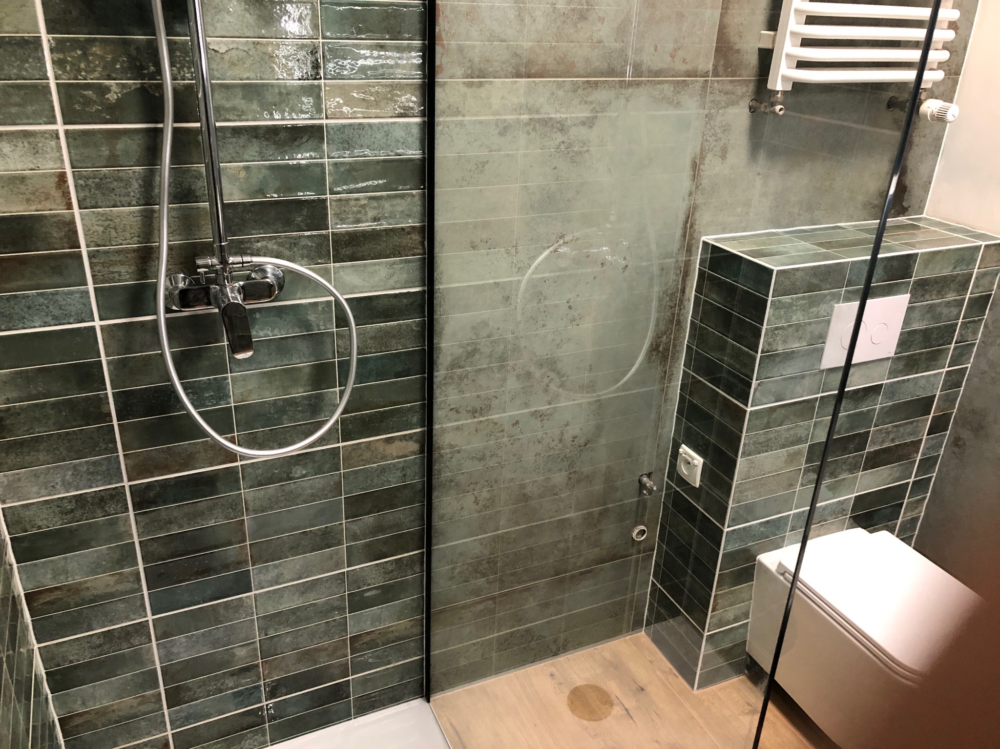 Nowoczesna kabina prysznicowa z armaturą w kolorze chromu i zielonymi, podłużnymi płytkami, uzupełniona o drewnopodobną podłogę i wiszącą toaletę z obudową z płytek w tym samym kolorze.