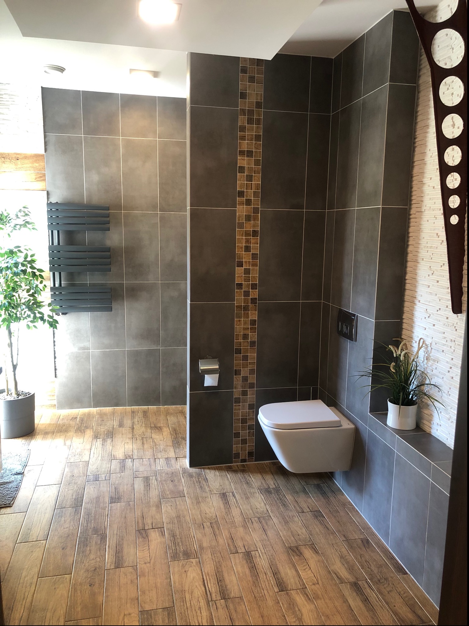 Nowoczesna łazienka z szarymi płytkami, drewnopodobną podłogą, wiszącą toaletą i dekoracyjną kolumną z mozaiki oraz nietypowym zegarem na ścianie.