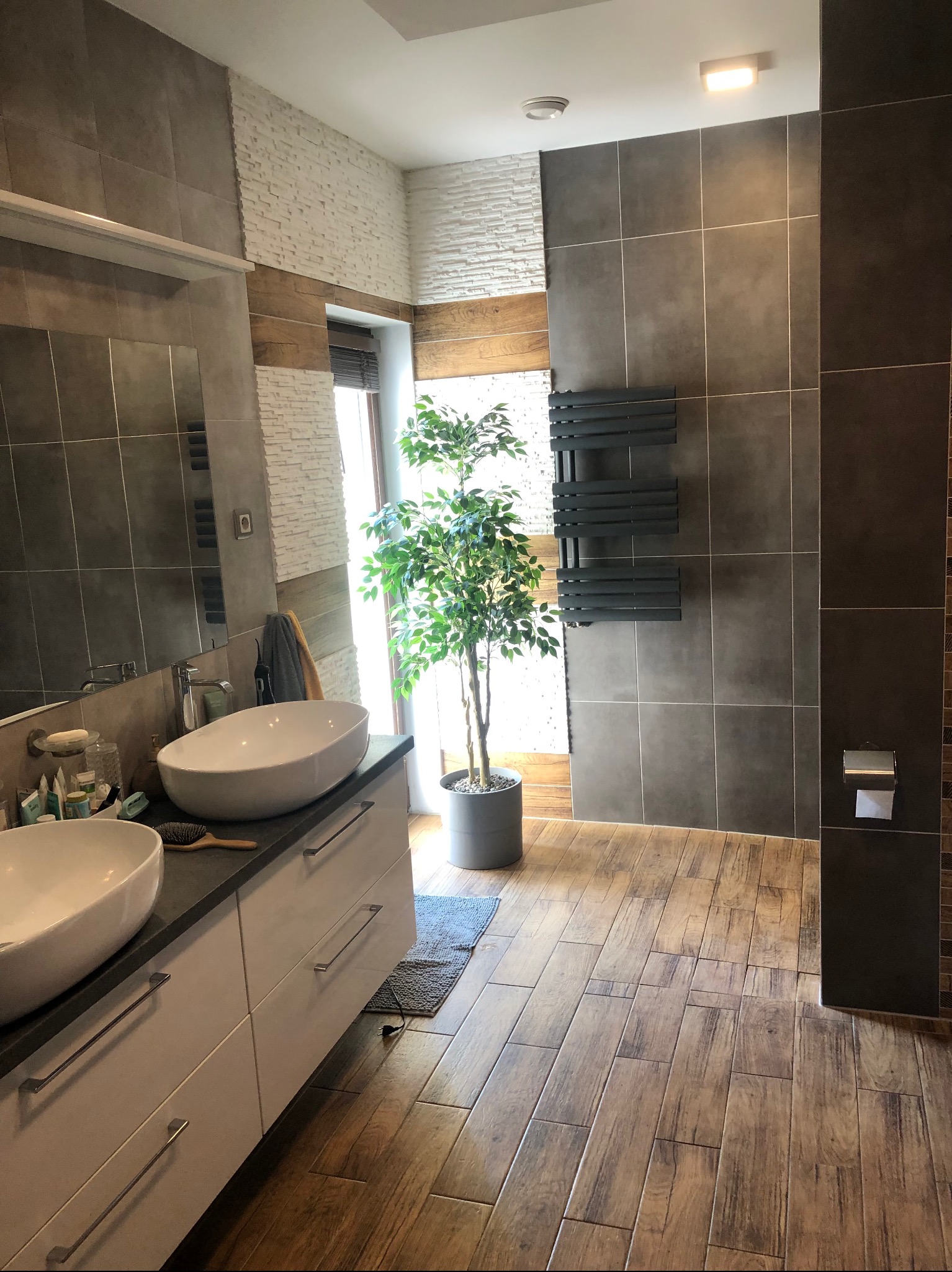 Nowoczesna łazienka z dwoma owalnymi umywalkami nablatowymi, szarą płytką imitującą kamień na ścianach, drewnopodobną podłogą, grzejnikiem drabinkowym i rośliną w doniczce.