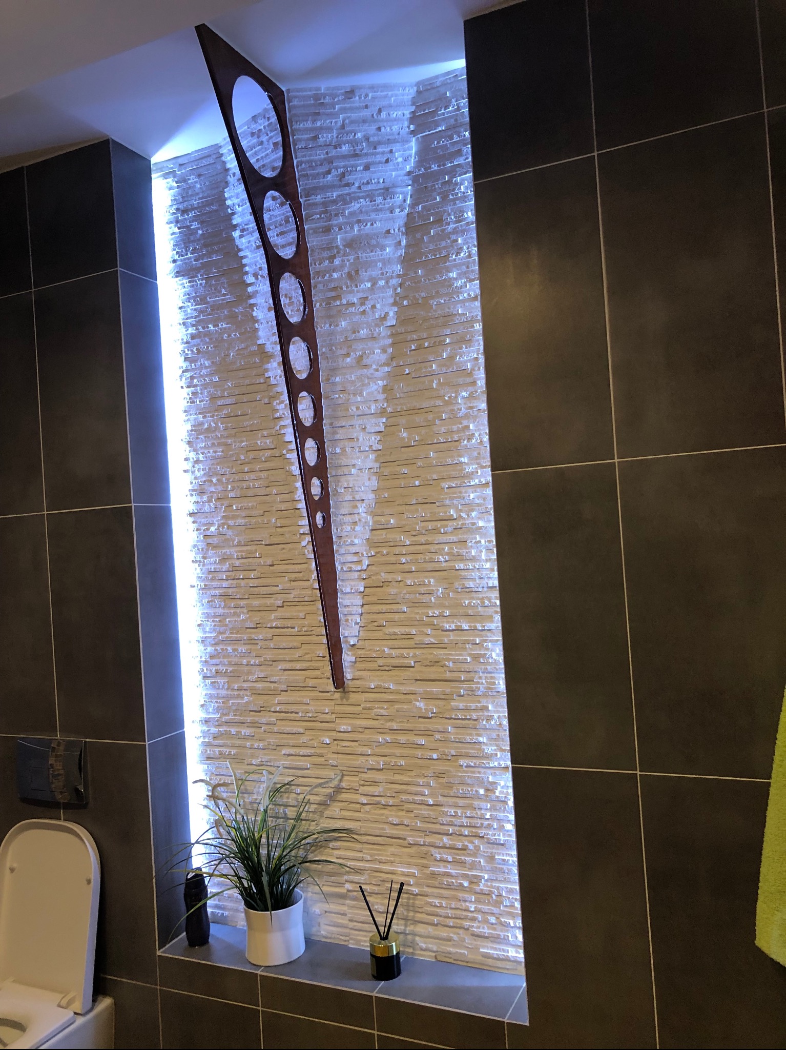 Nowoczesna łazienka z podświetloną ścianą z dekoracyjnej cegły, drewnianą ozdobą z wyciętymi okręgami, rośliną w białej doniczce i czarnymi, kwadratowymi płytkami na ścianach.