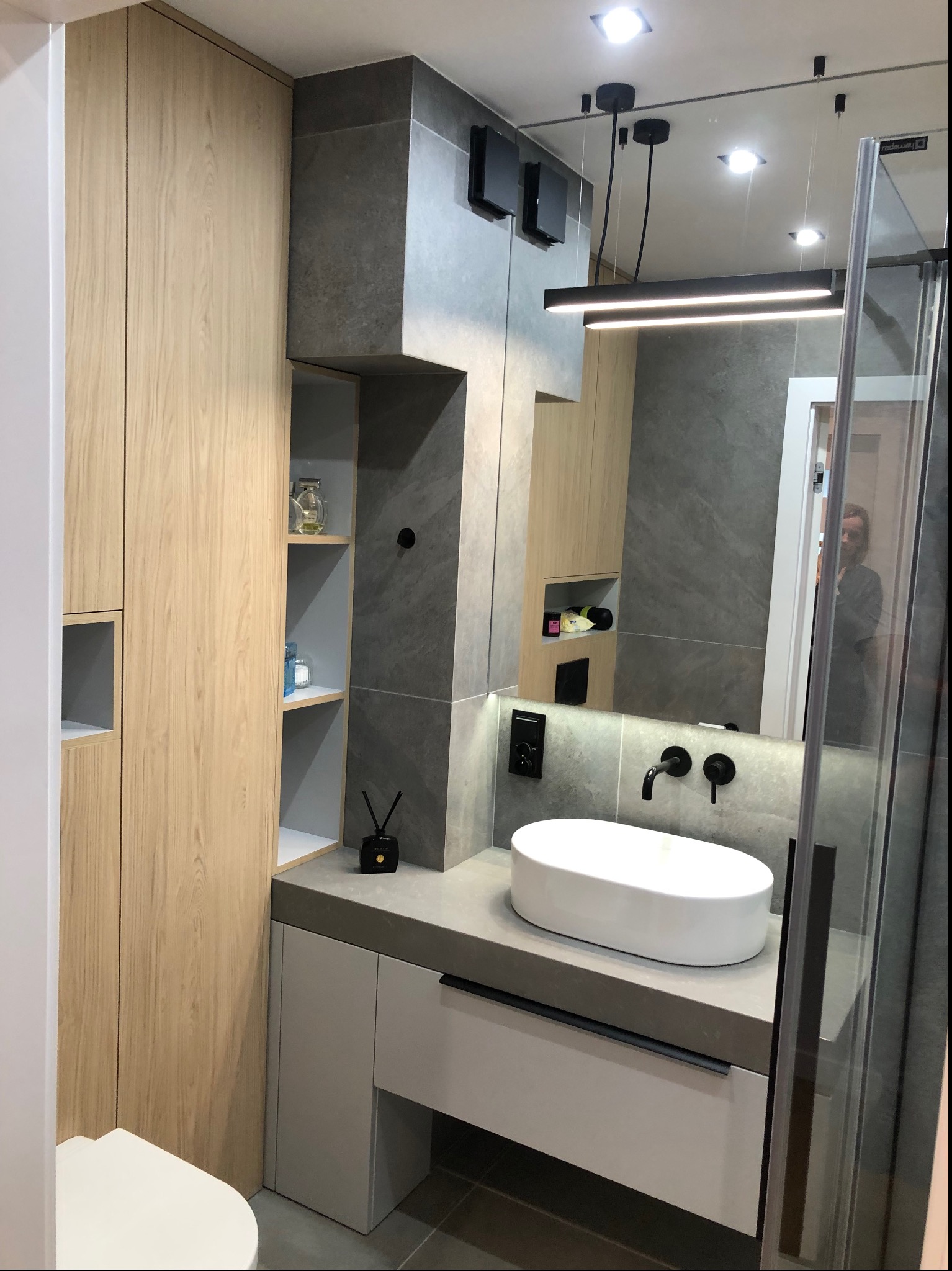 Nowoczesna łazienka z drewnianą szafką, szarą umywalką nablatową i czarną armaturą, oświetlona podłużną lampą LED, z widocznym fragmentem kabiny prysznicowej.