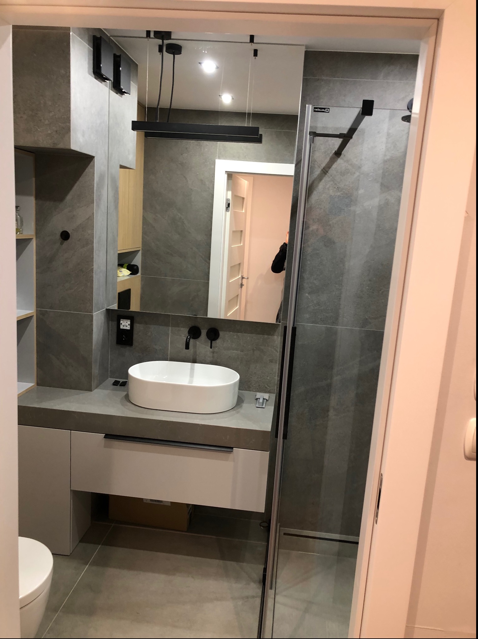 Nowoczesna łazienka z umywalką nablatową, szarą płytką imitującą kamień, czarną armaturą i minimalistycznym oświetleniem; widoczna kabina prysznicowa typu walk-in i fragment toalety.