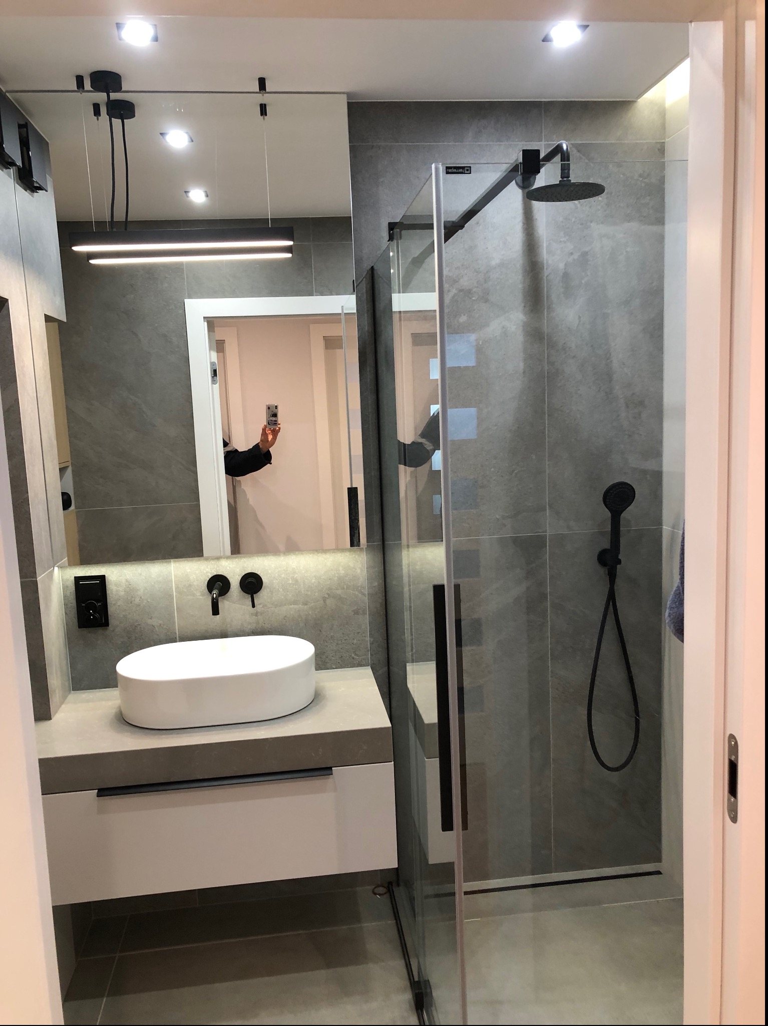 Nowoczesna łazienka z szarymi płytkami, kabiną prysznicową typu walk-in z czarną armaturą, owalną białą umywalką nablatową i podświetlanym lustrem, w którym odbija się osoba robiąca zdjęcie telefonem.