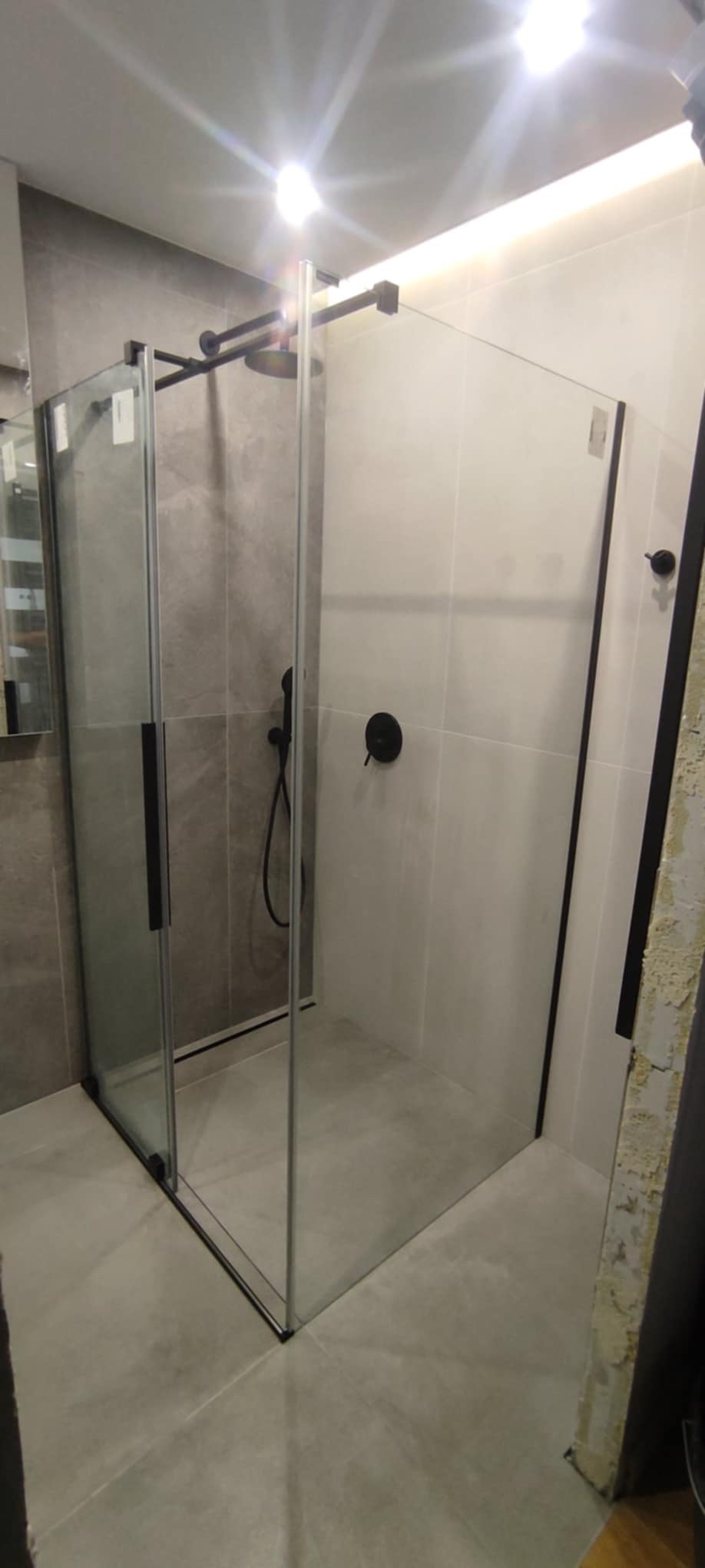 Nowoczesna kabina prysznicowa z czarnymi elementami wykończeniowymi, zamontowana w łazience z szarymi i białymi płytkami, widoczna deszczownica i bateria podtynkowa.