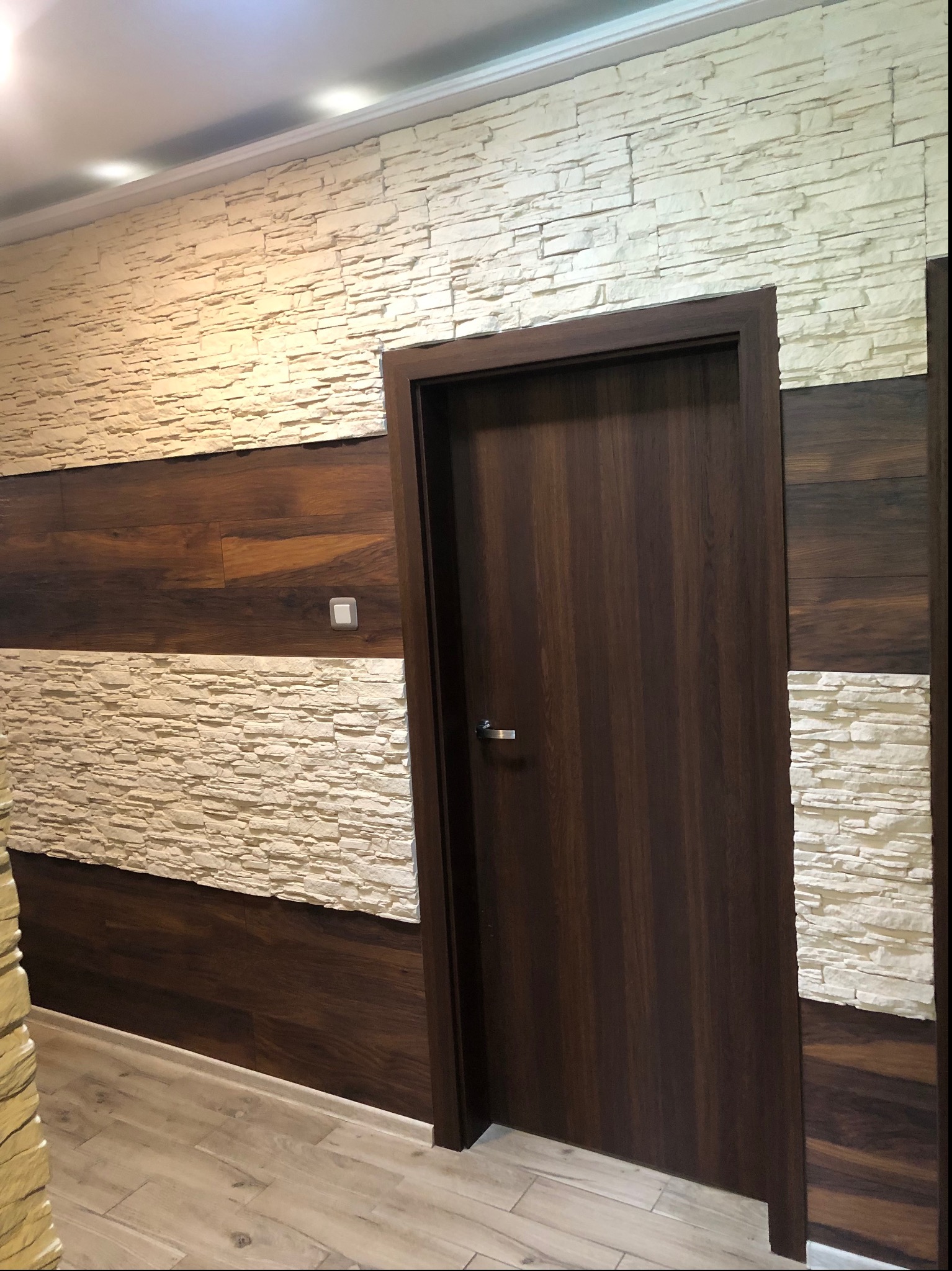 Wnętrze z dekoracyjną ścianą z imitacji kamienia w odcieniach beżu, przedzieloną poziomym pasem z paneli drewnopodobnych w kolorze ciemnego brązu, oraz z ciemnymi drzwiami z metalową klamką.