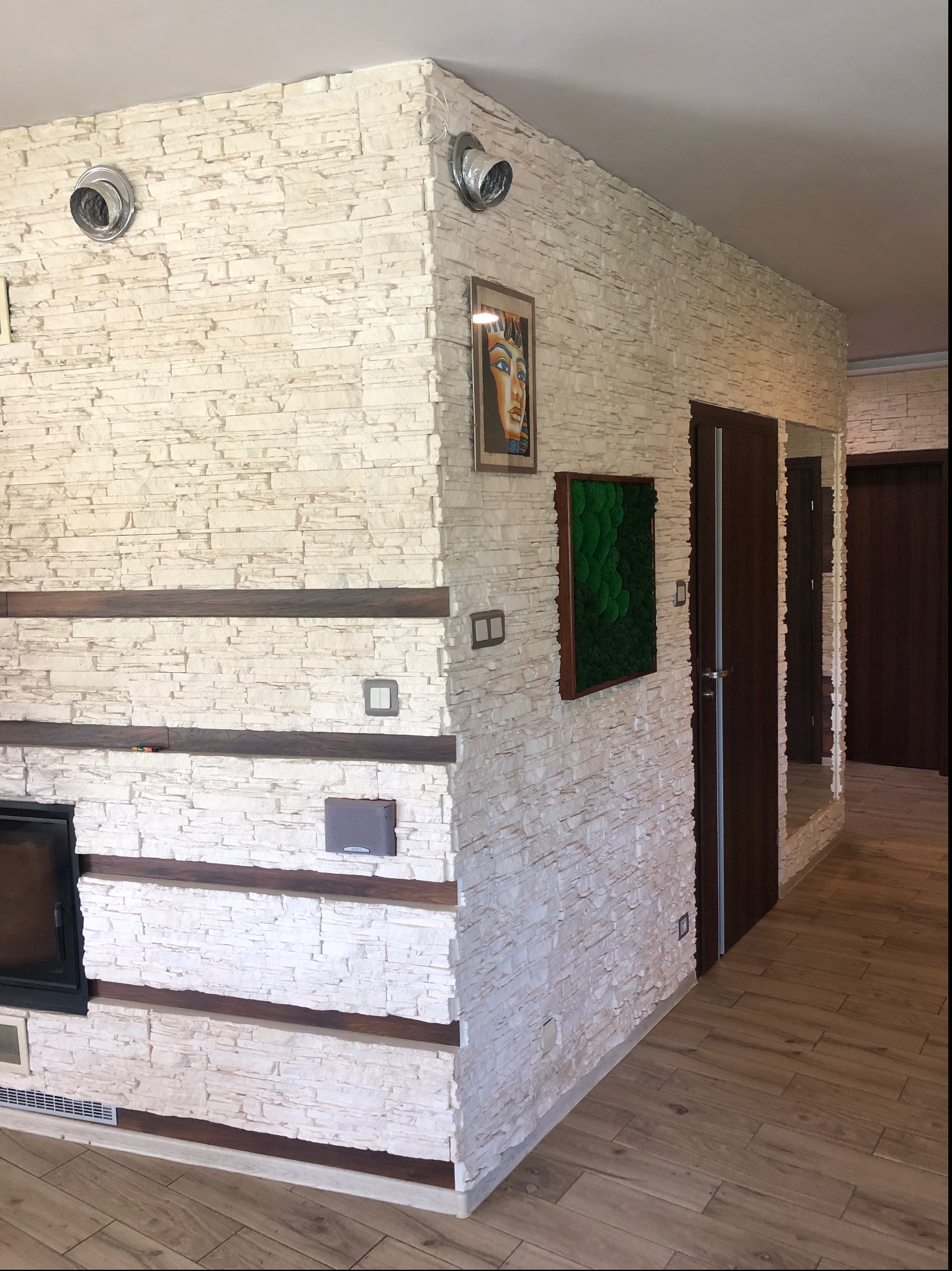 Fragment wykończonego wnętrza z dekoracyjną ścianą z białego kamienia, drewnianymi elementami, obrazem z motywem egipskim, panelem z mchu i drewnianą podłogą.