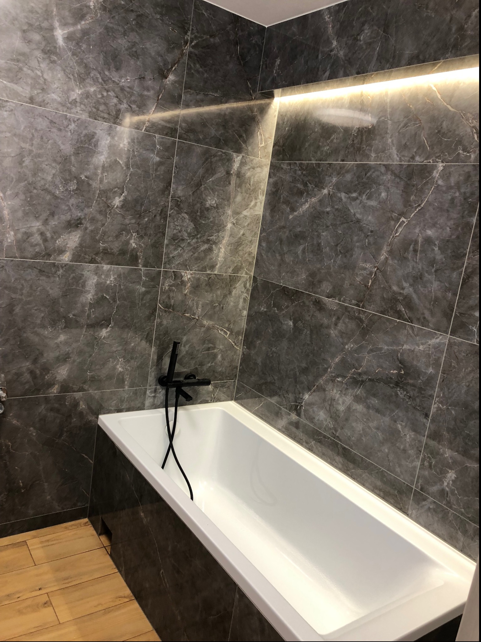 Biała wanna z czarną armaturą na tle ściany wyłożonej płytkami imitującymi ciemny marmur, dodatkowo oświetlona listwą LED.