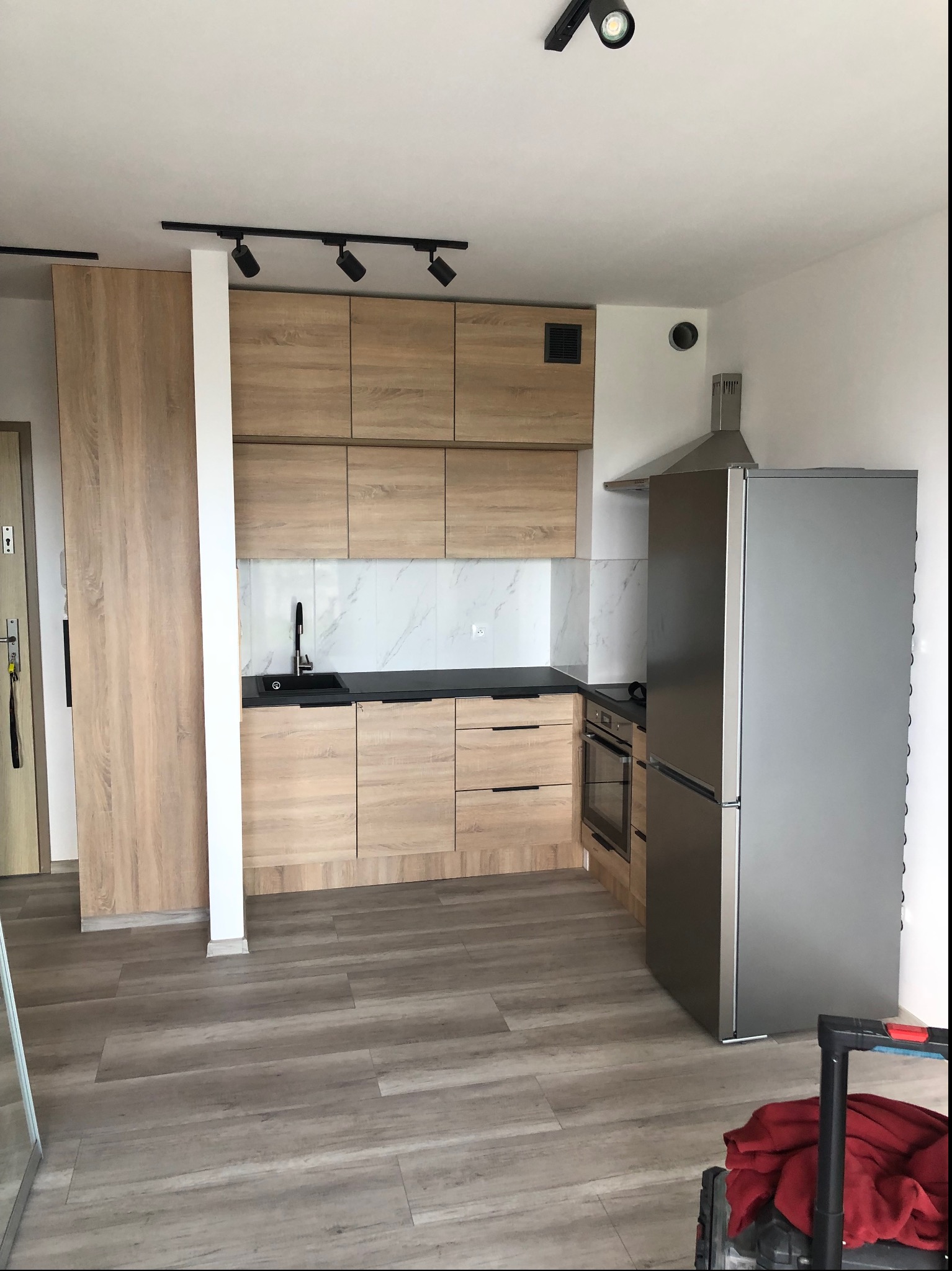 Nowoczesna kuchnia z drewnianymi szafkami, czarnym blatem i marmurowym backsplashiem, oświetlona reflektorami sufitowymi, z widoczną lodówką i fragmentem podłogi z paneli imitujących drewno.