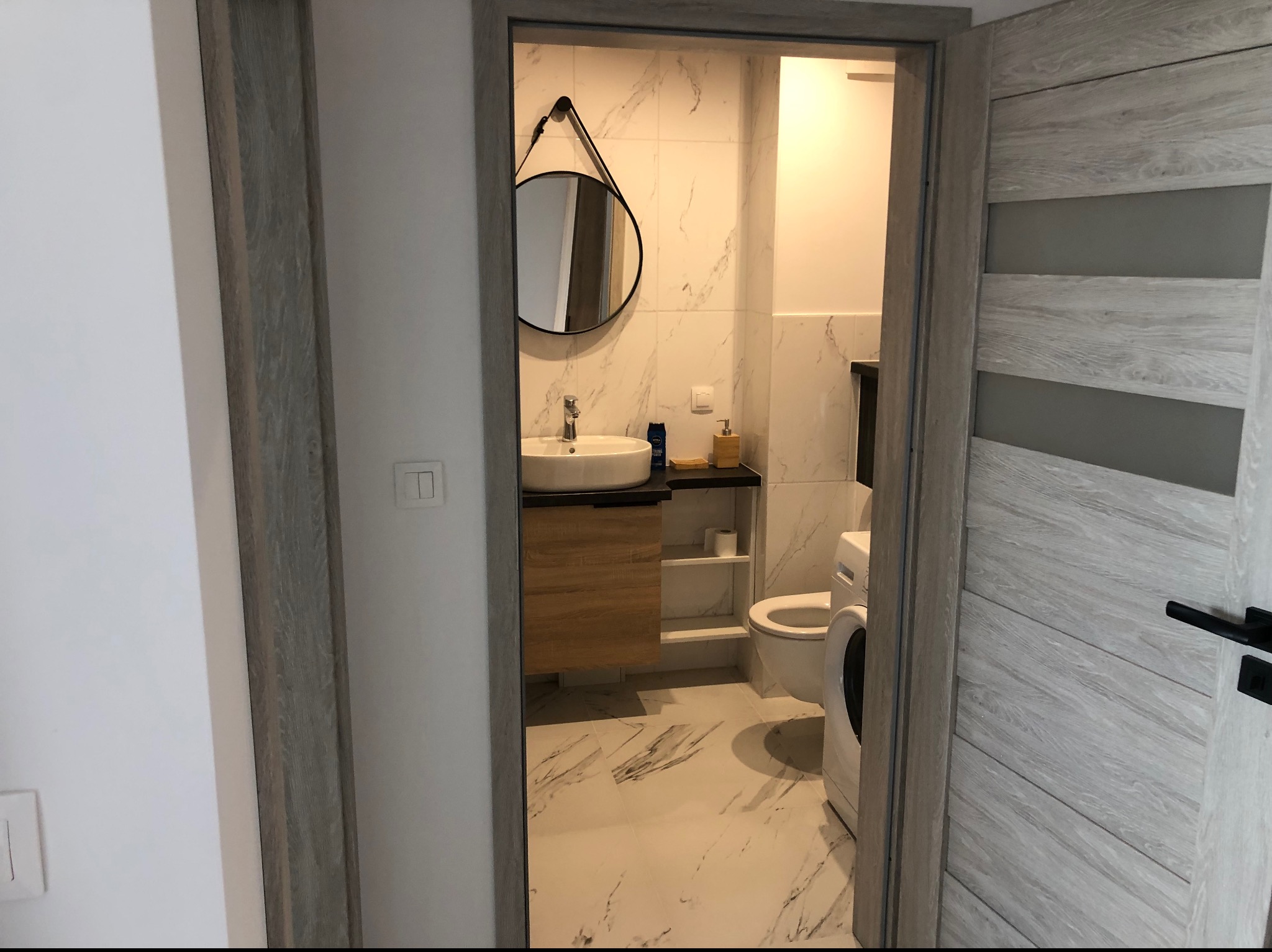 Widok na nowoczesną łazienkę z umywalką na drewnianej szafce, okrągłym lustrem na marmurowej ścianie i pralką, widzianą zza otwartych drzwi o teksturze drewna.