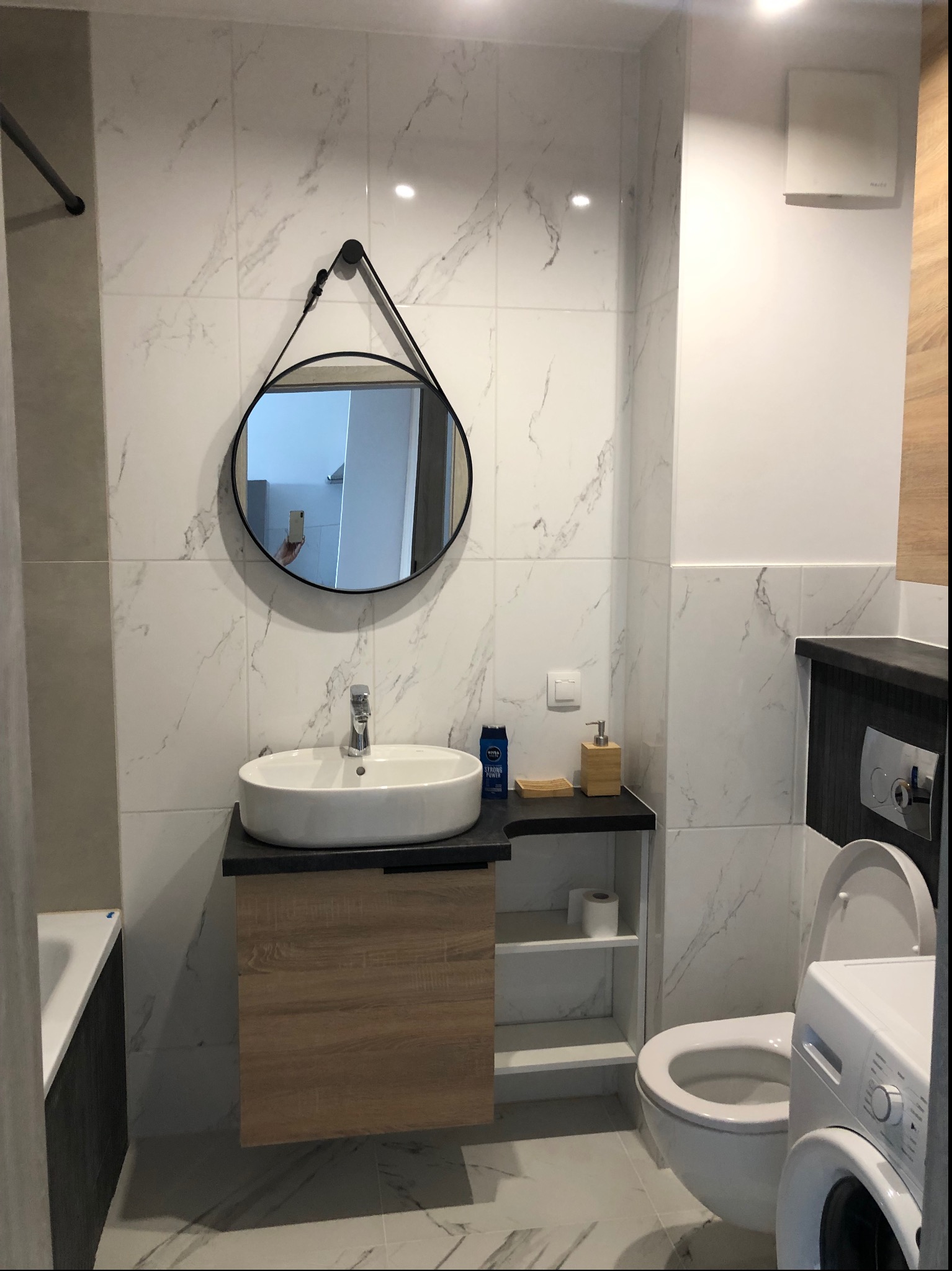 Nowoczesna łazienka z marmurowymi płytkami, owalną umywalką na szafce z drewnopodobnym frontem, okrągłym lustrem na pasku i pralką obok toalety.