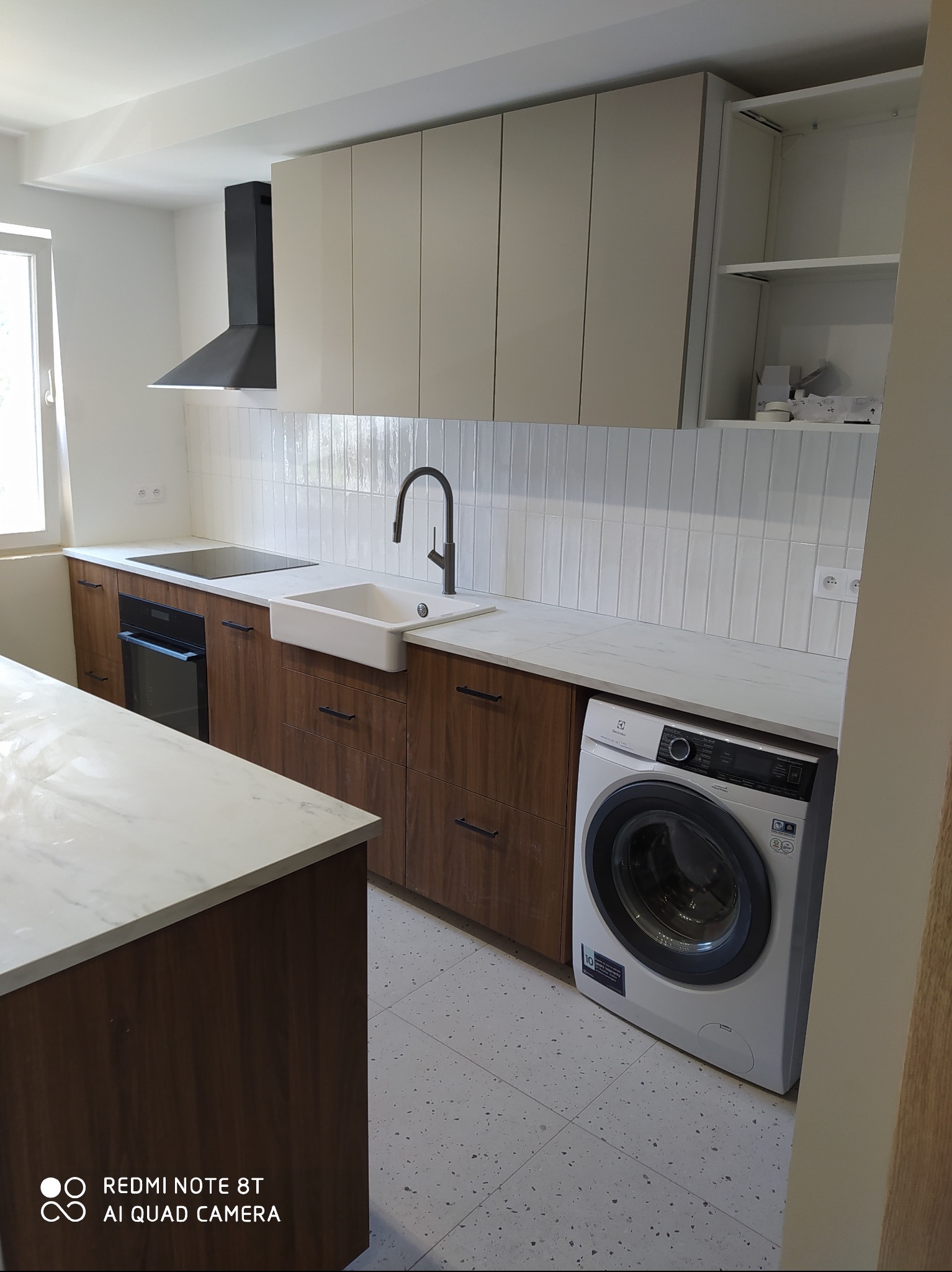 Nowoczesna kuchnia z drewnianymi szafkami, białym blatem imitującym marmur, płytkami wertykalnymi nad blatem i pralką w zabudowie, widok na fragment wyspy kuchennej.