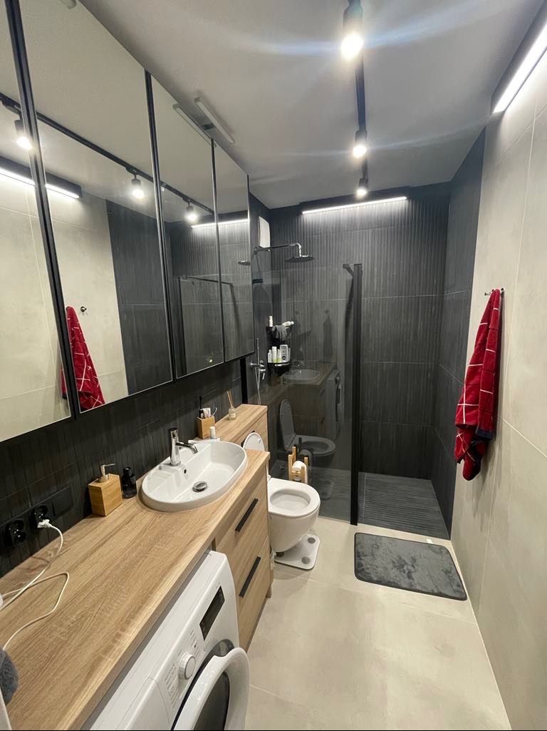 Nowoczesna łazienka z kabiną prysznicową typu walk-in, umywalką nablatową na drewnianym blacie, pralką pod blatem, dużymi lustrami w czarnych ramach i ciemnymi płytkami na ścianach.