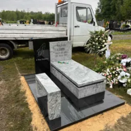 Czarny nagrobek z jasnoszarym nakryciem i ławką, ozdobiony grawerem rakiety i napisem '# Pan Torpeda', ustawiony na cmentarzu, obok dekoracje kwiatowe i samochód dostawczy.